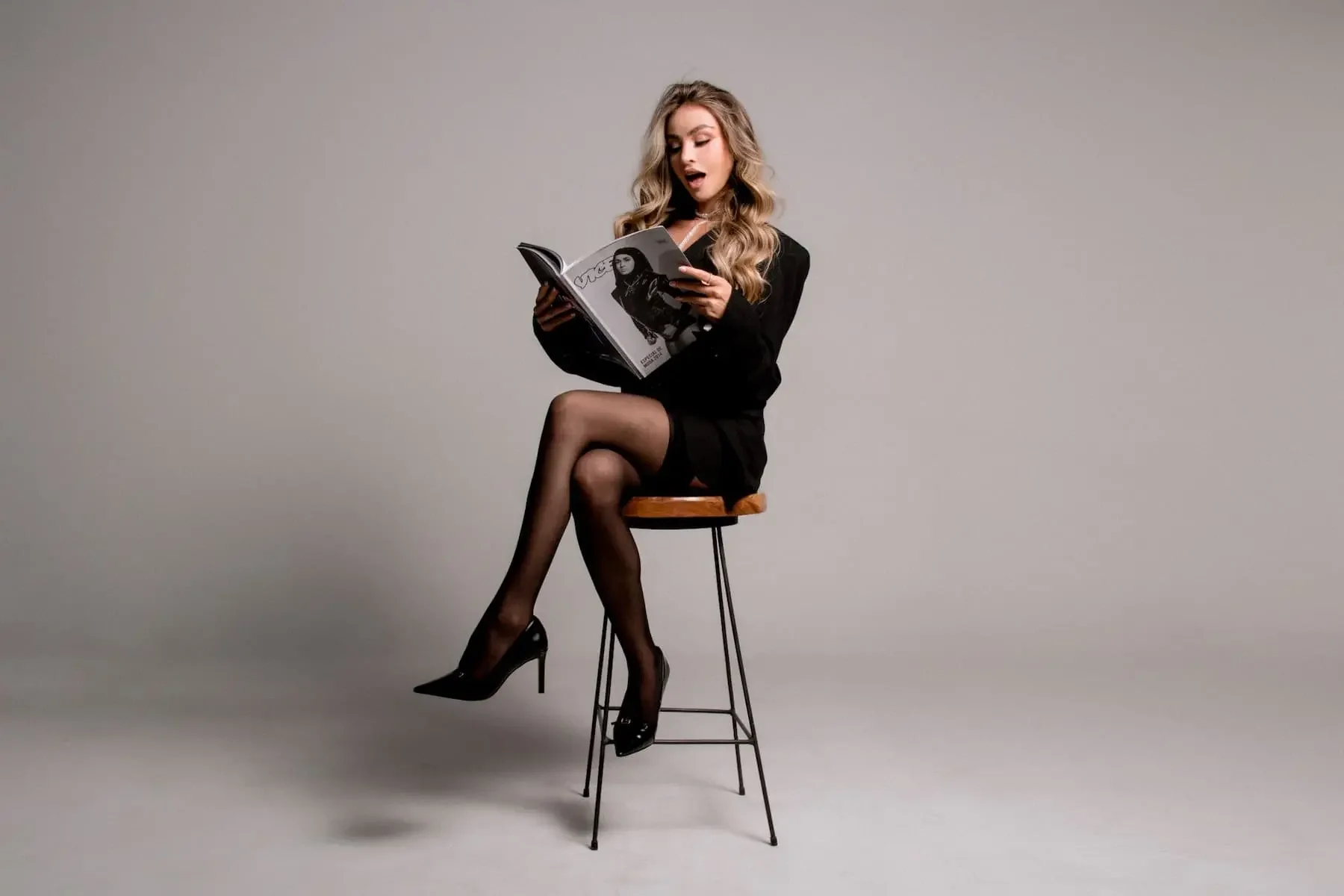 Mujer joven con cabello rubio y ondulado, usando conjunto negro y tacones negros, sentada en un taburete de madera leyendo una revista con una imagen de otra mujer en la portada, fondo gris liso.