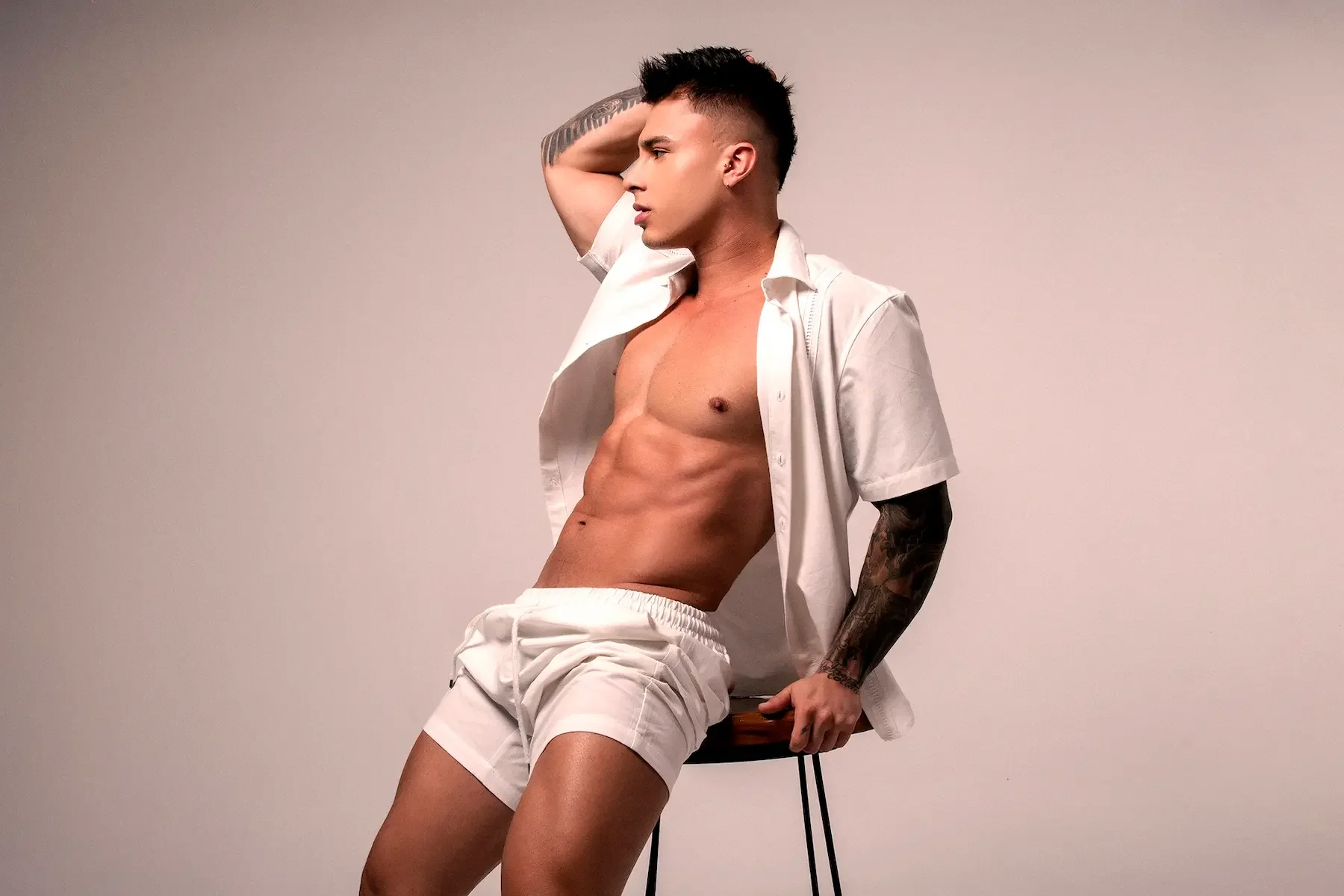 Joven hombre de perfil en ropa ligera, sentado en una silla, con brazo en la cabeza y camisa abierta, mostrando abdominales y tatuajes.