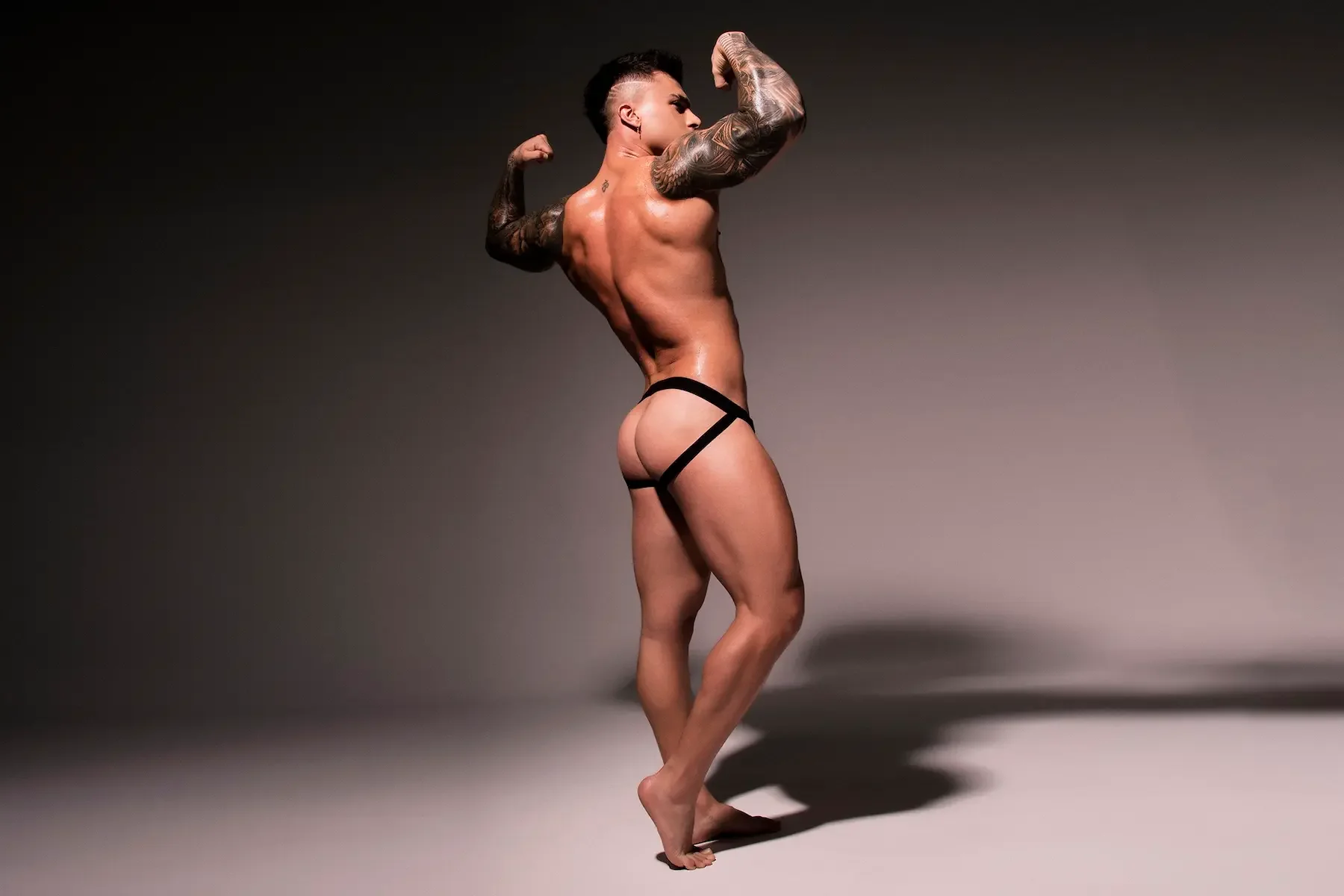 Hombre musculoso en calzón negro, posando con los músculos flexionados, en un fondo gris, mostrando su físico.