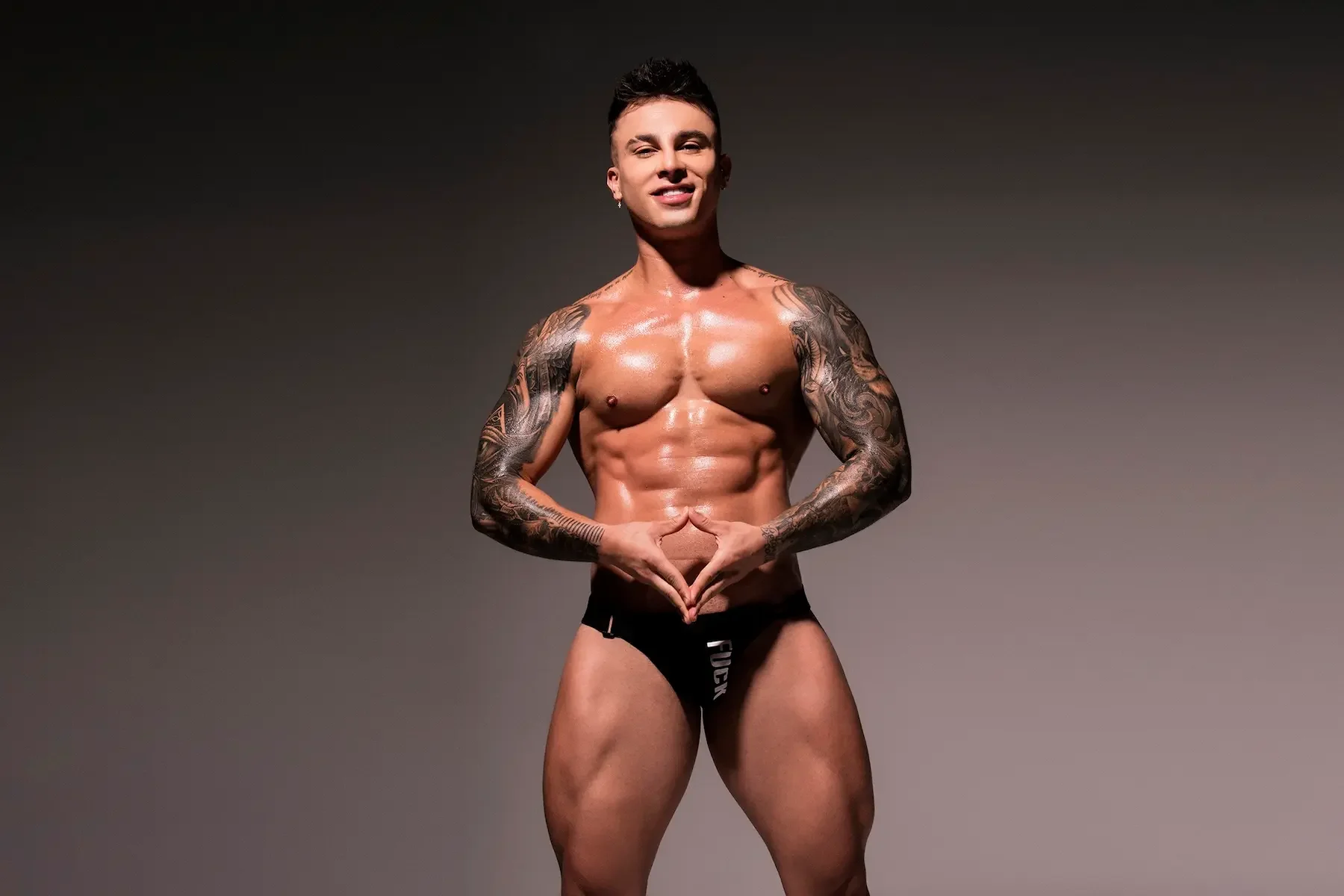 Hombre musculoso con tatuajes, posando en ropa interior negra, haciendo signo de corazón con las manos, contra fondo gris.