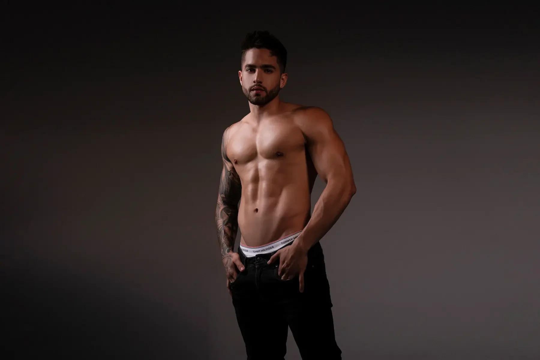 Joven con torso desnudo y pantalones oscuros, con tatuajes en un brazo, frente a fondo oscuro.