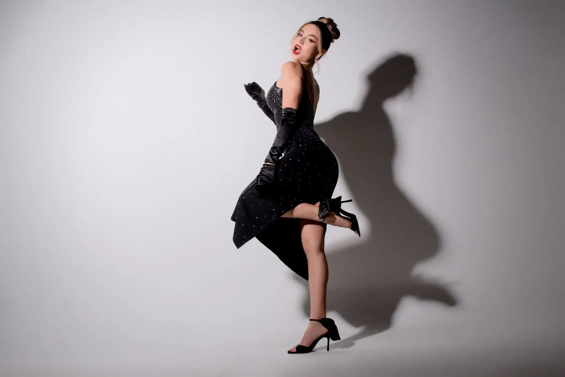 Mujer con vestido negro, tacones y guantes largos, posando en contra de una pared blanca con sombra visible.