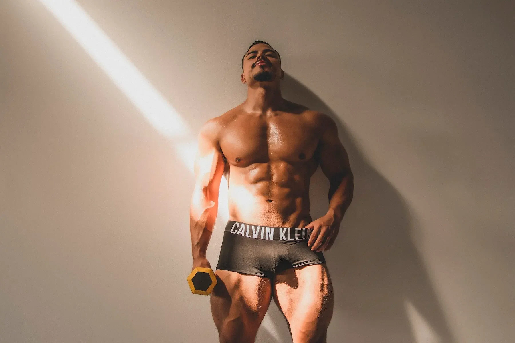 Hombre musculoso con pantalones cortos Calvin Klein, sosteniendo una pesa hexagonal amarilla, de pie frente a una pared y con iluminación que crea su sombra en la pared.