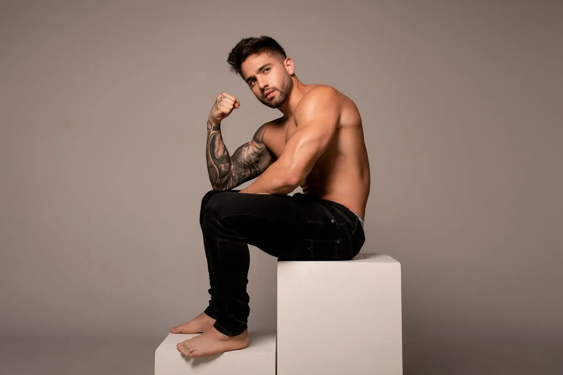 Joven hombre sin camisa, con tatuajes en el brazo izquierdo, sentado en un bloque blanco, haciendo un gesto de fuerza con el puño, fondo gris.