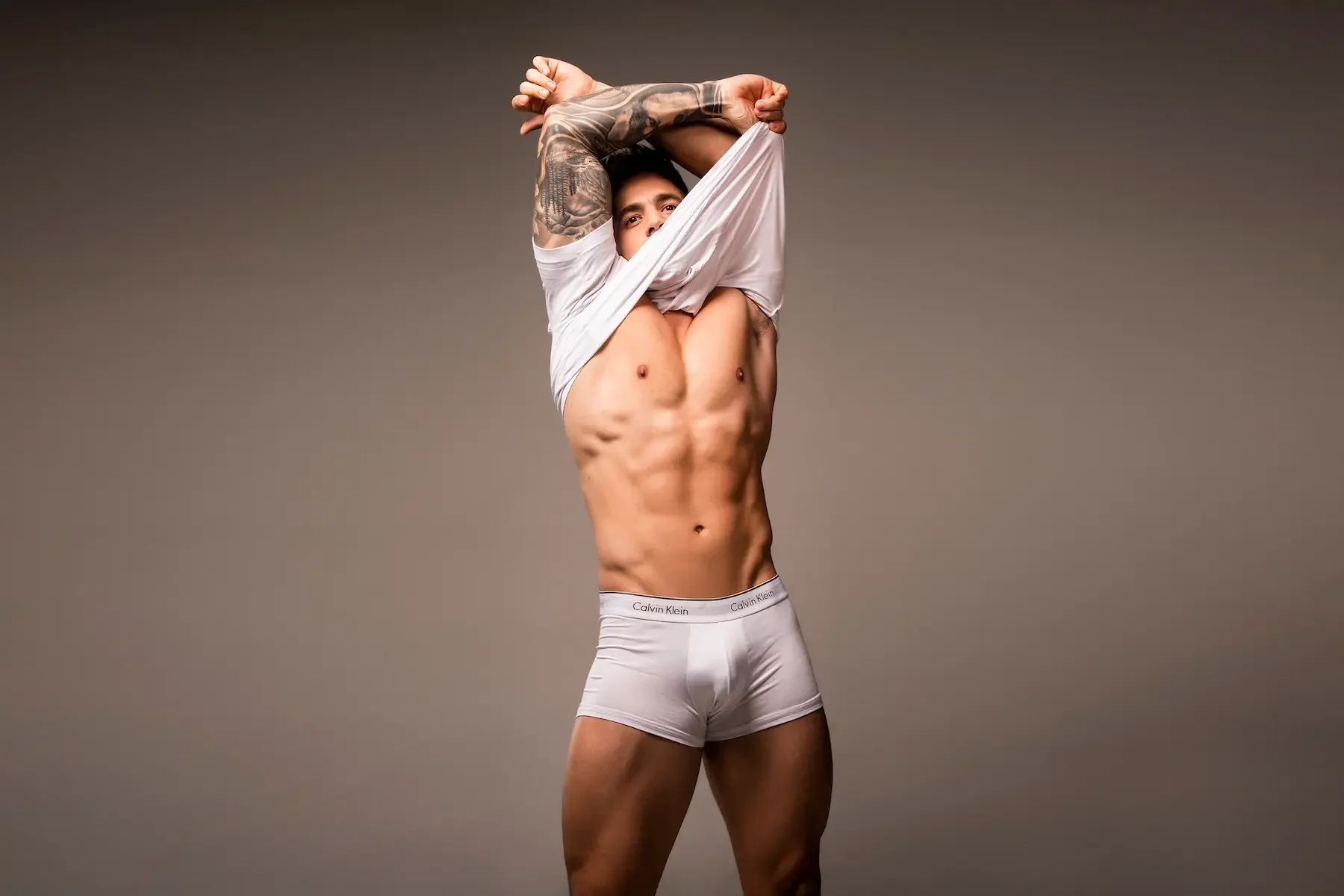 Hombre musculoso con tatuajes, levantando una camiseta blanca y usando ropa interior blanca de la marca Calvin Klein, en un fondo gris liso.