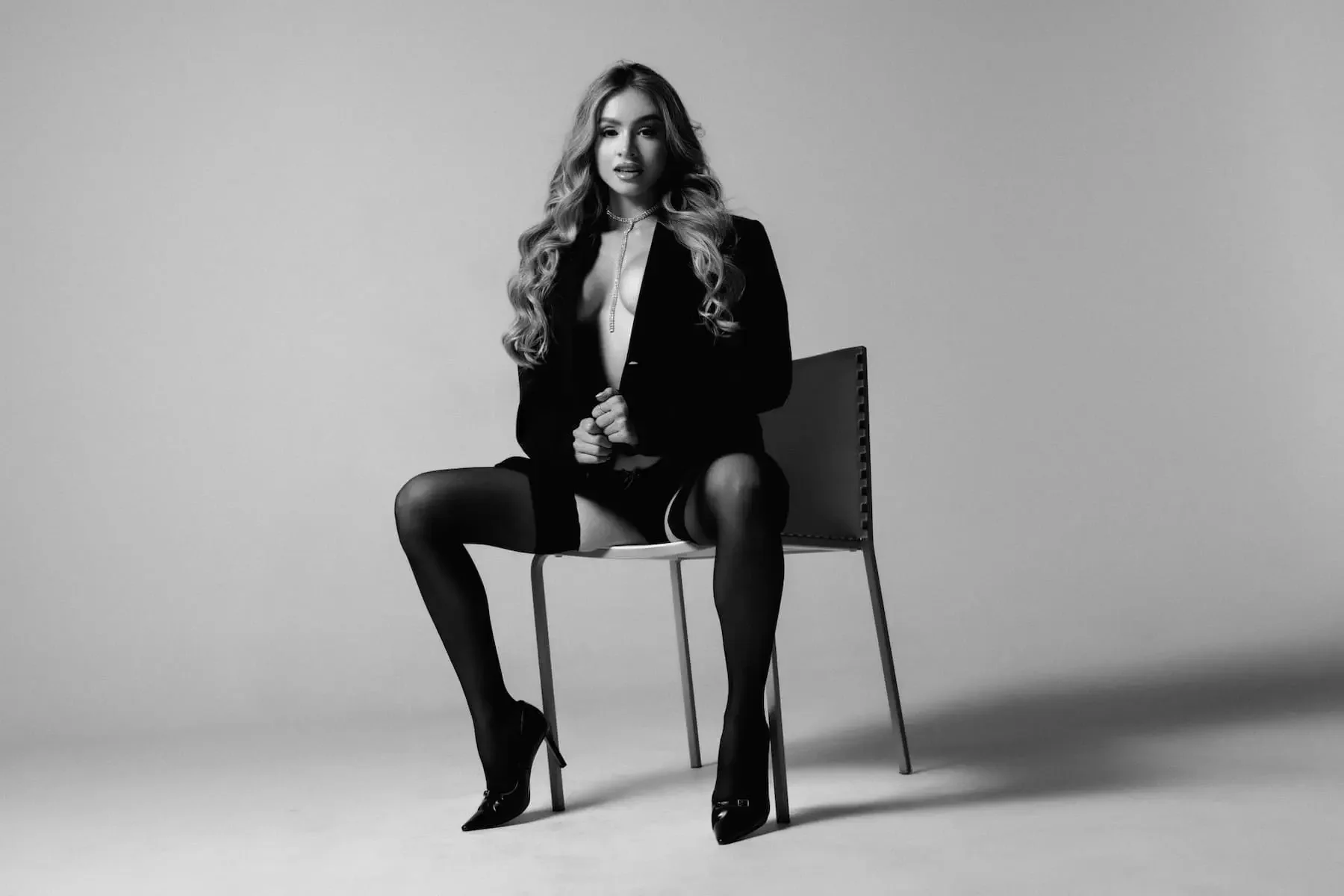Mujer con cabello rizado largo, vestida con traje negro y tacones, sentada en una silla en un fondo gris, en una foto en blanco y negro.