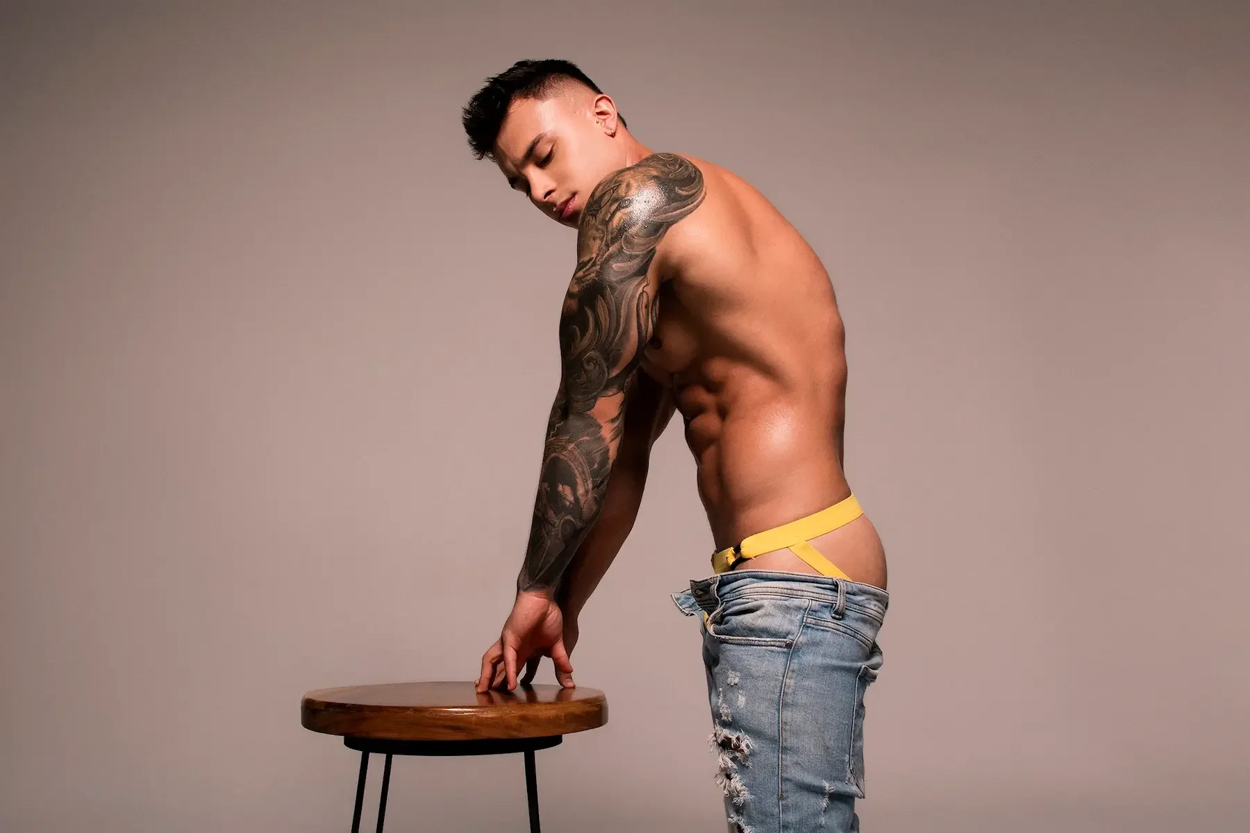 Hombre musculoso con tatuajes, sin camisa, sosteniendo una mesa de madera y usando pantalones rotos.