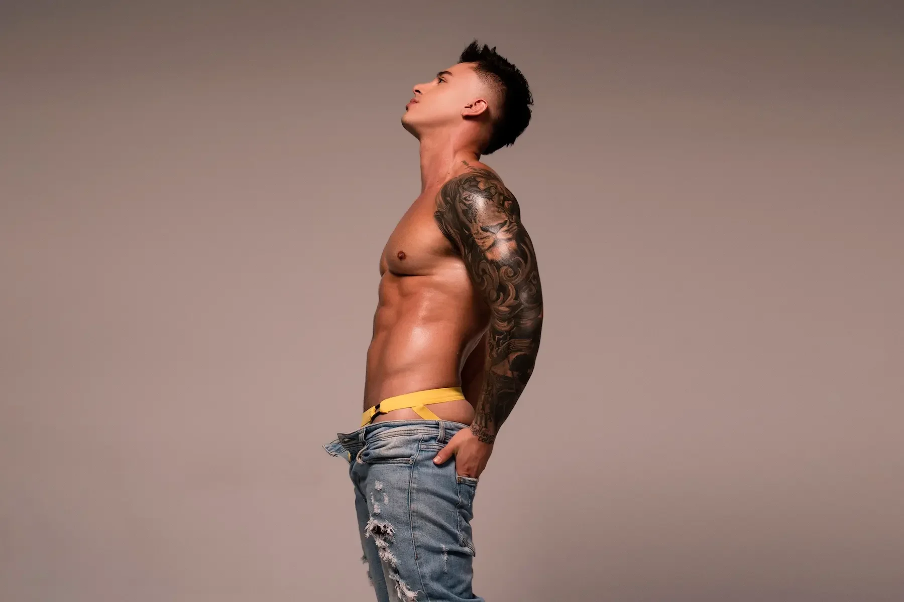 Joven hombre de lado sin camisa, con tatuaje en el brazo, con pantalones desgastados y sin camiseta, posando en estudio con fondo neutro.