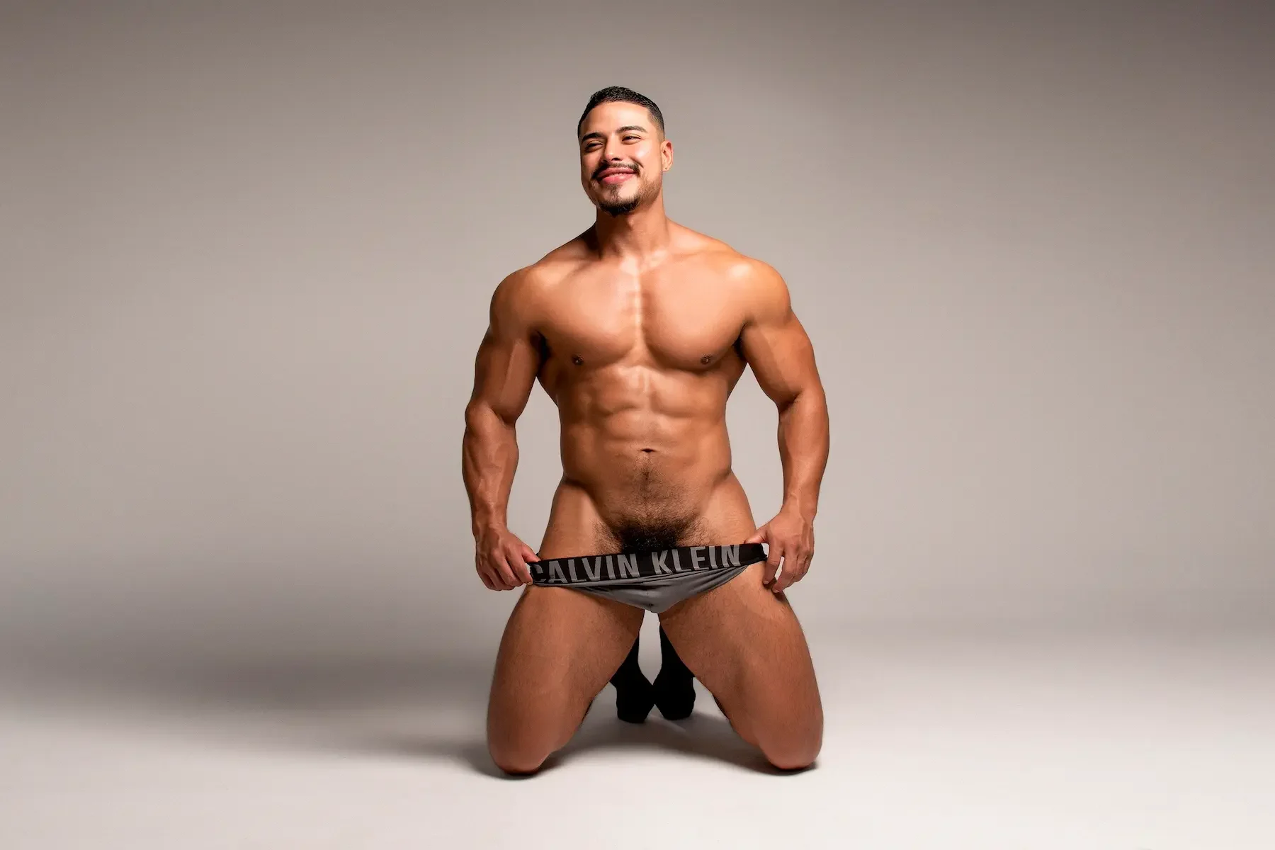 Hombre musculoso, desnudo, en posición de rodillas, sosteniendo cinturilla de calzón Calvin Klein, sonrisa, fondo neutro.