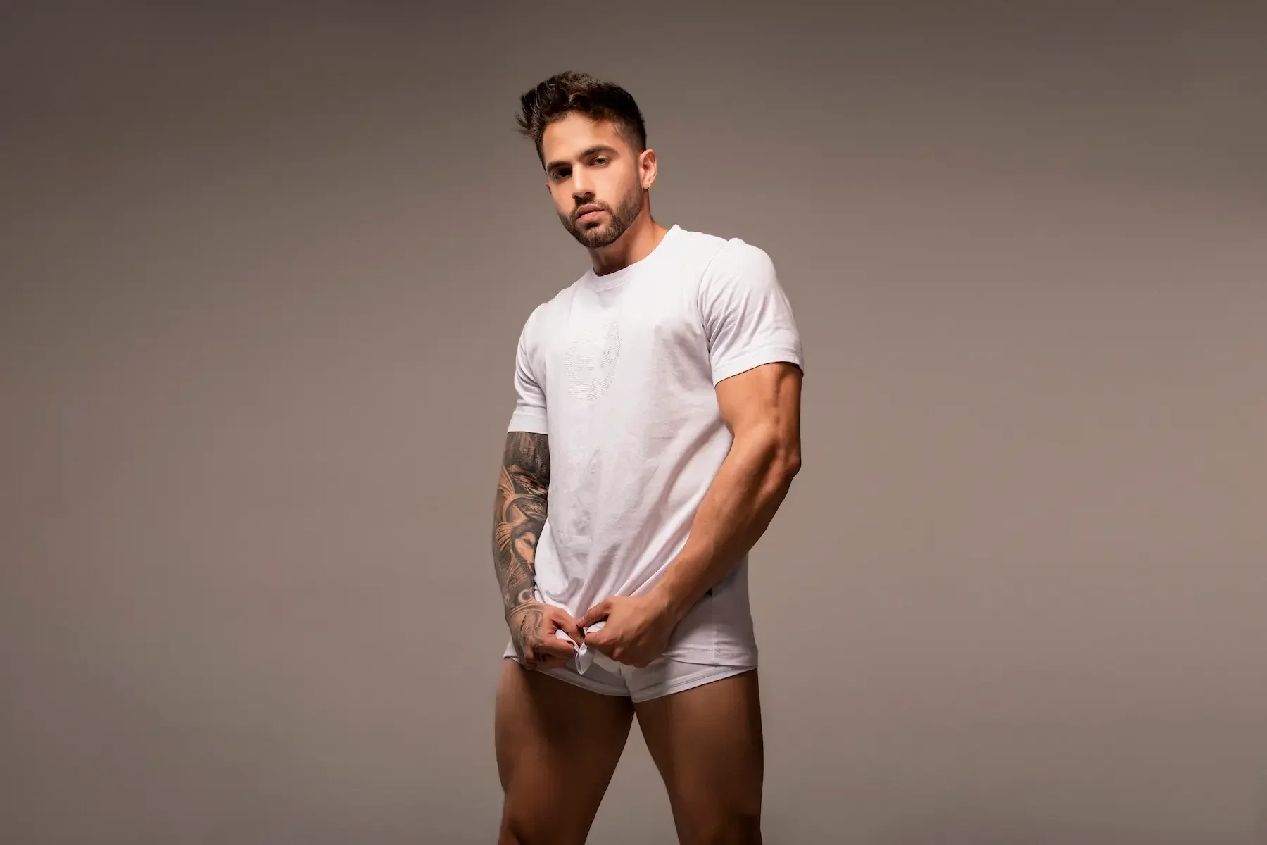 Joven con barba y tatuajes en el brazo izquierdo, usando camiseta blanca y ropa interior blanca, posando con expresión seria frente a fondo gris.
