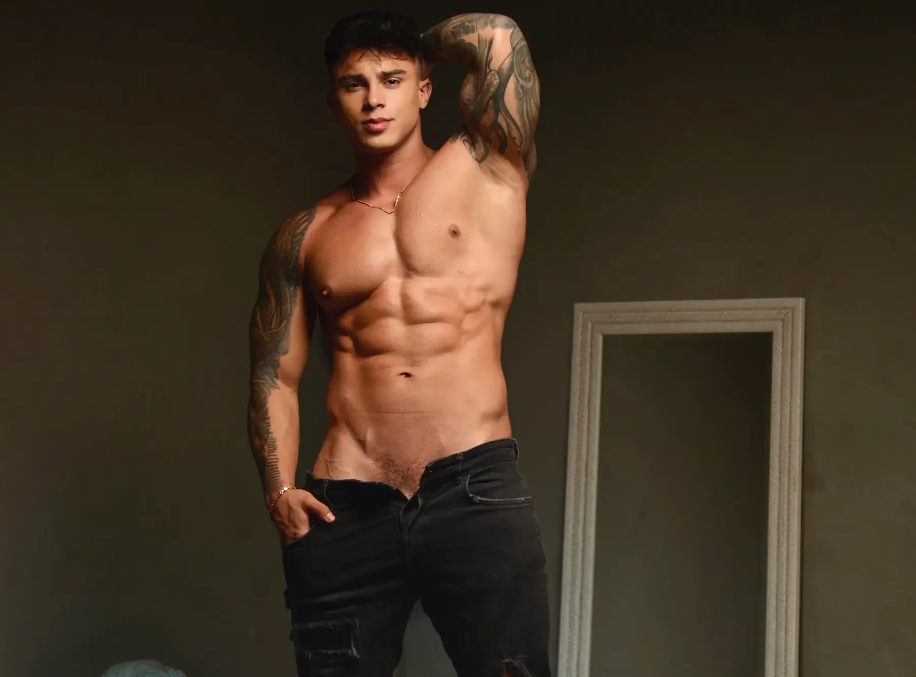 Joven sin camiseta con tatuajes en los brazos, posando con una mano detrás de la cabeza en un cuarto con espejo.