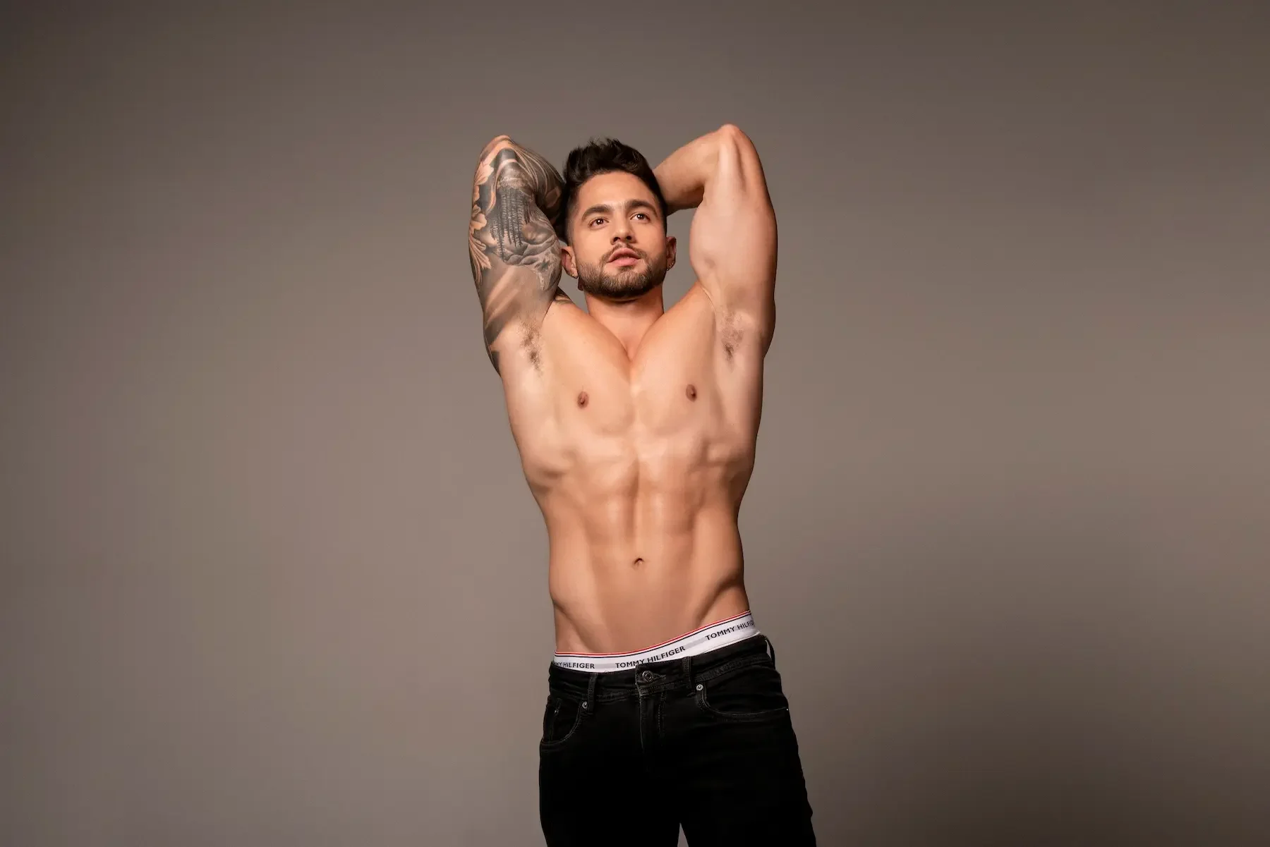 Joven hombre musculoso con tatuajes en un brazo, en jeans y sin camiseta, posando con las manos detrás de la cabeza frente a un fondo gris.