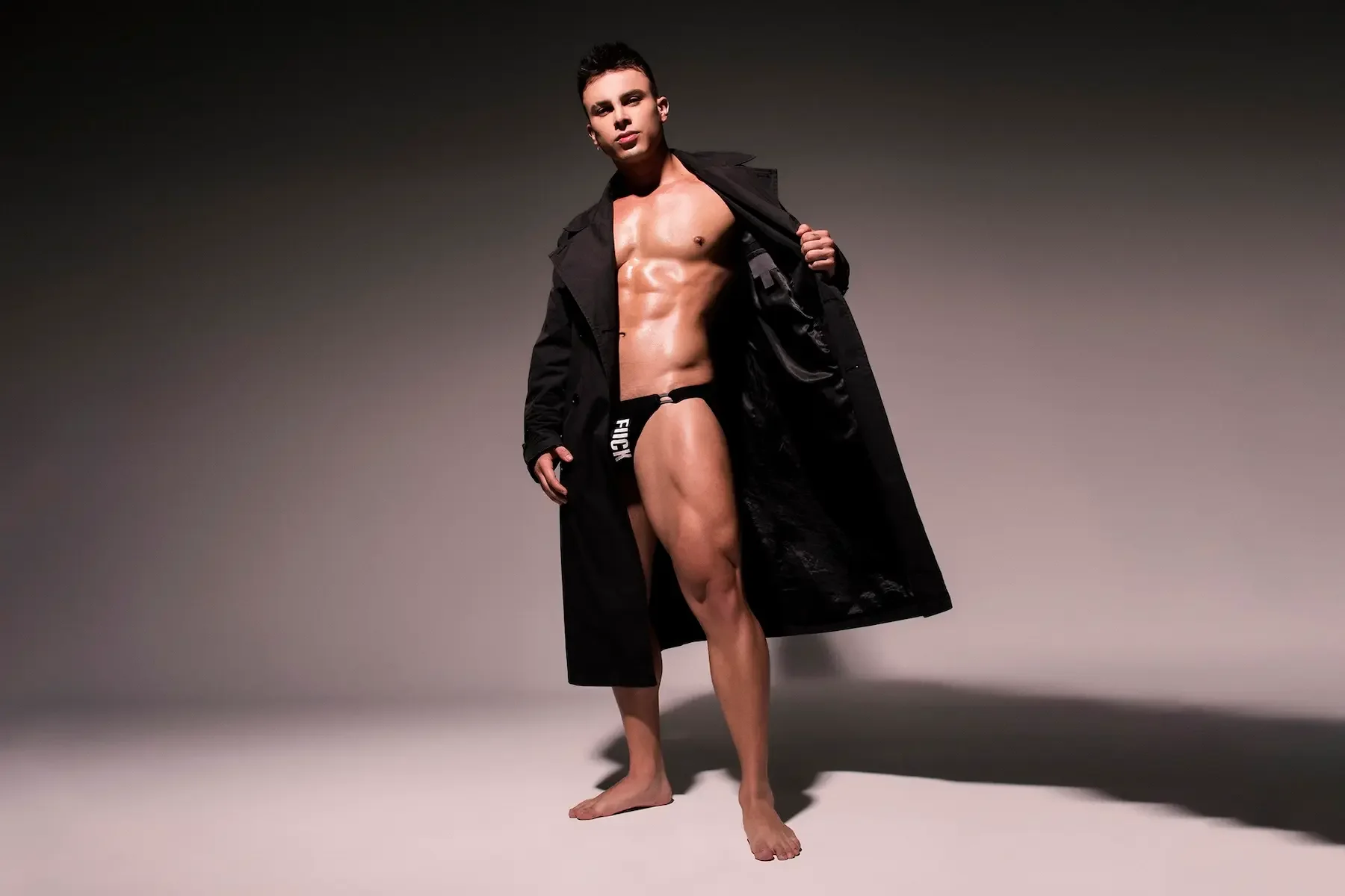 Hombre con cuerpo musculoso y aspecto masculino, usando solo un tanga negro y una gabardina negra, en un fondo gris oscuro.
