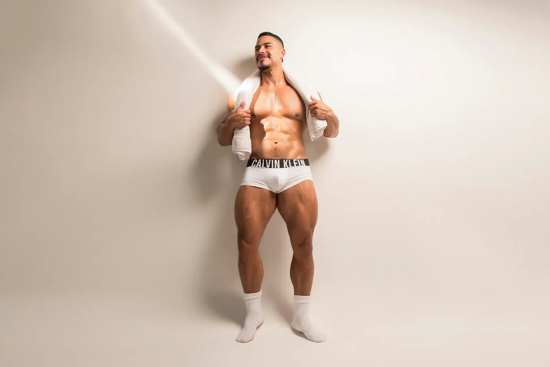 Hombre musculoso con toalla, usando calzoncillos blancos Calvin Klein, sonriendo con fondo blanco.