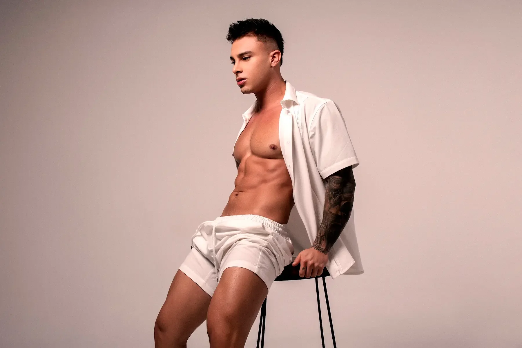Hombre joven con pantalones cortos y camisa blanca abierta, sentado en una silla, mostrando abdomen y brazo tatuado en fondo gris claro.