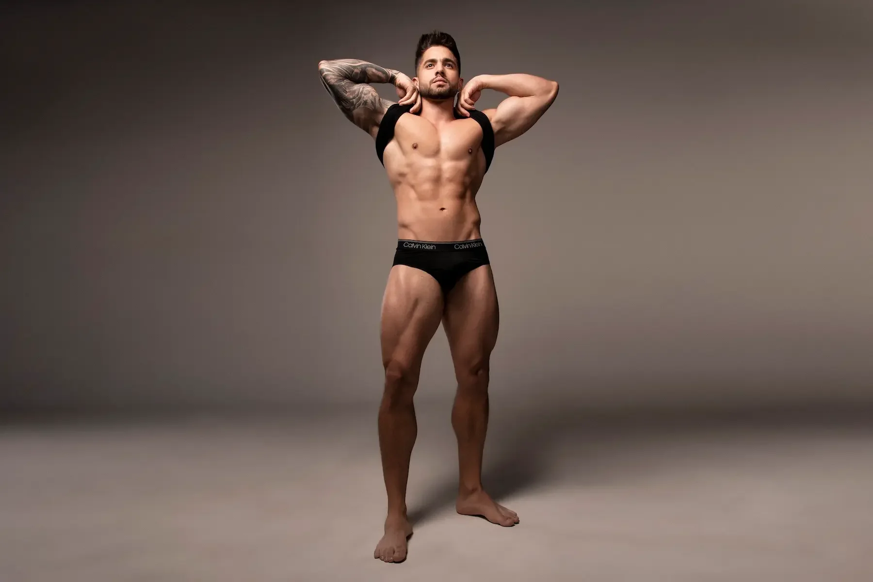 Hombre musculoso con tatuajes en brazos, usando ropa interior negra Calvin Klein, posando con manos detrás de la cabeza en un fondo neutro.