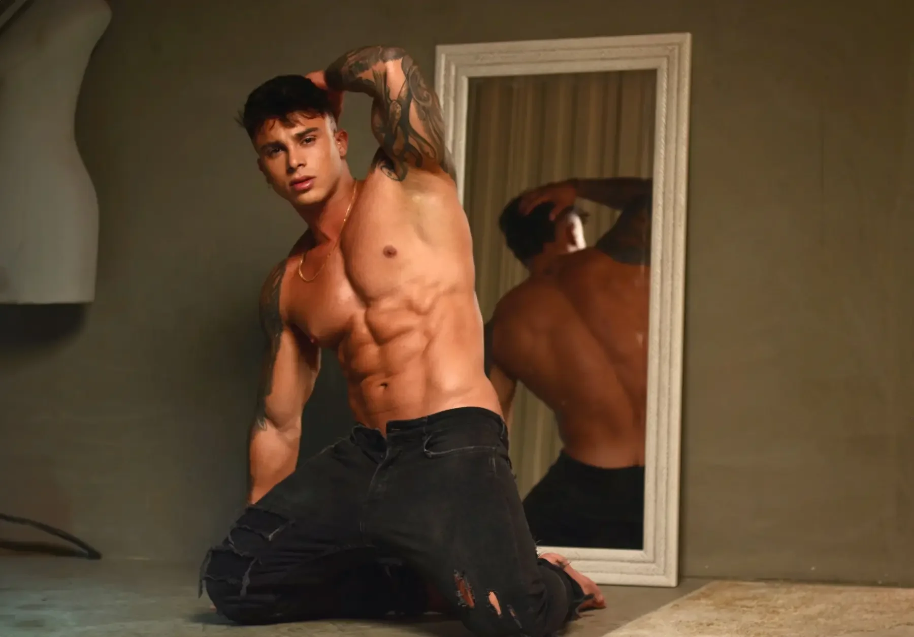Joven hombre con músculos, tatuajes, sin camisa, con pantalones oscuros, en cuclillas frente a un espejo, en un ambiente interior.
