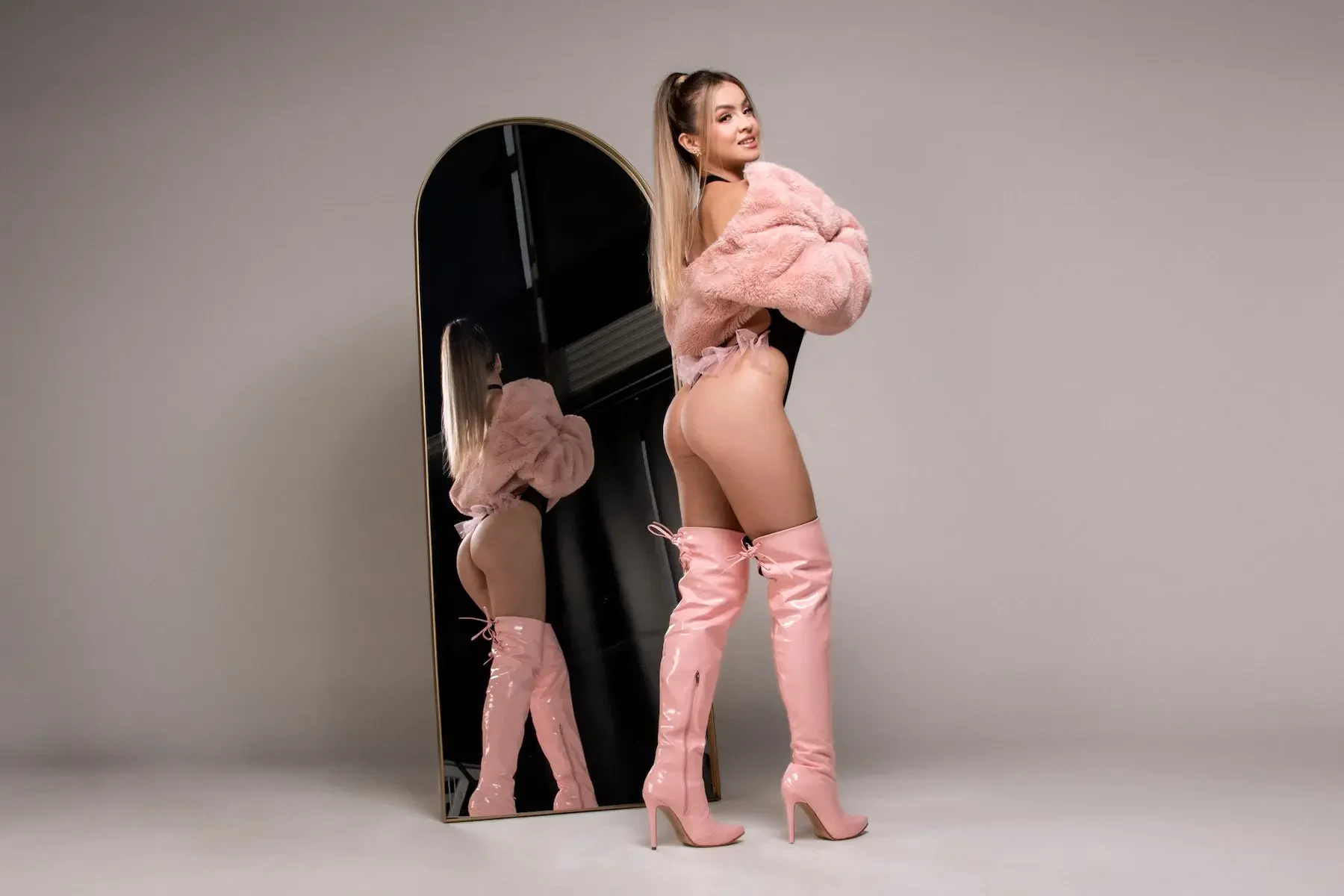 Joven mujer con botas altas rosadas y abrigo de peluche rosa frente a un espejo, sonriendo y posando.