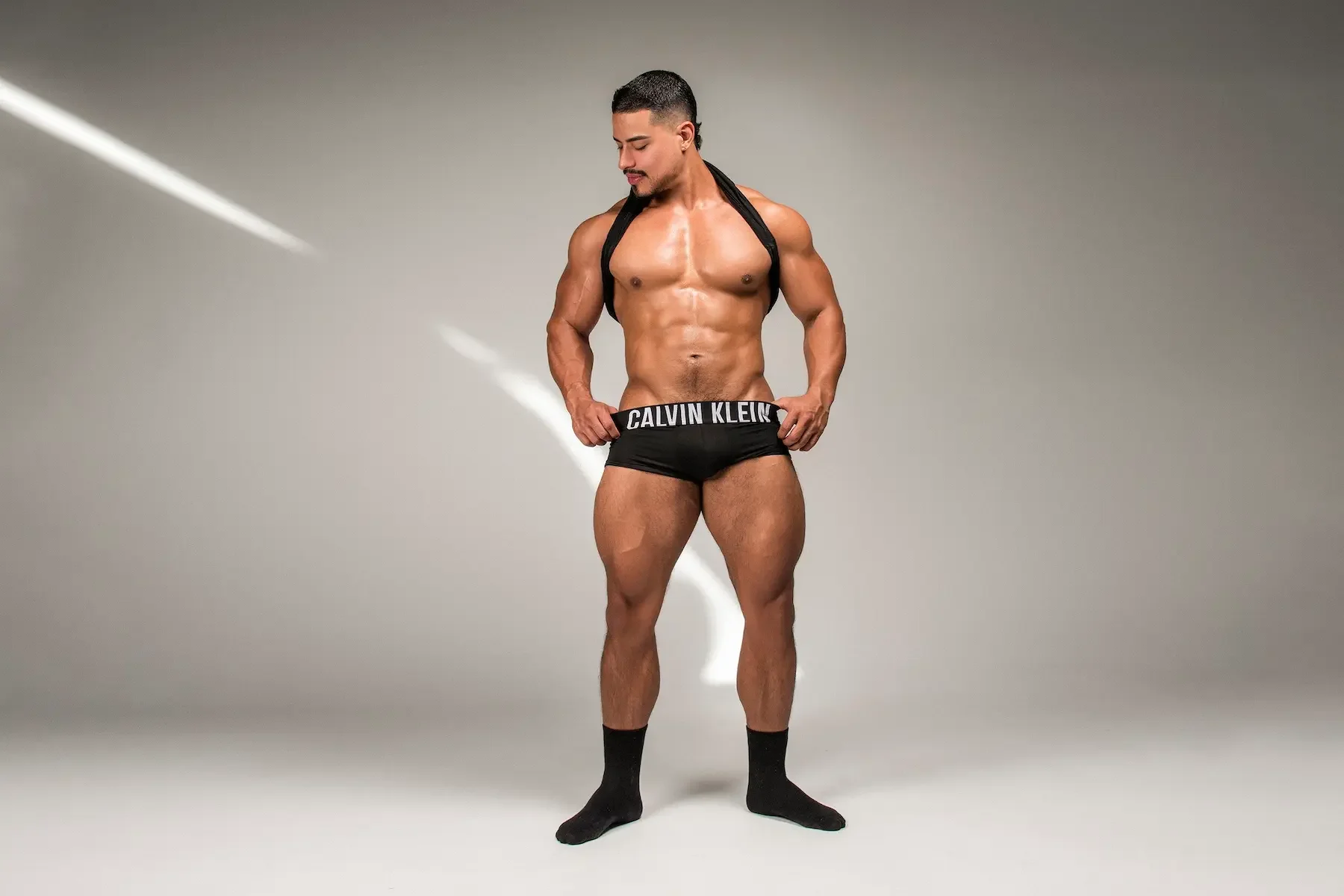 Hombre musculoso con short de Calvin Klein, con un saco colgado en los hombros, de pie en un fondo gris.