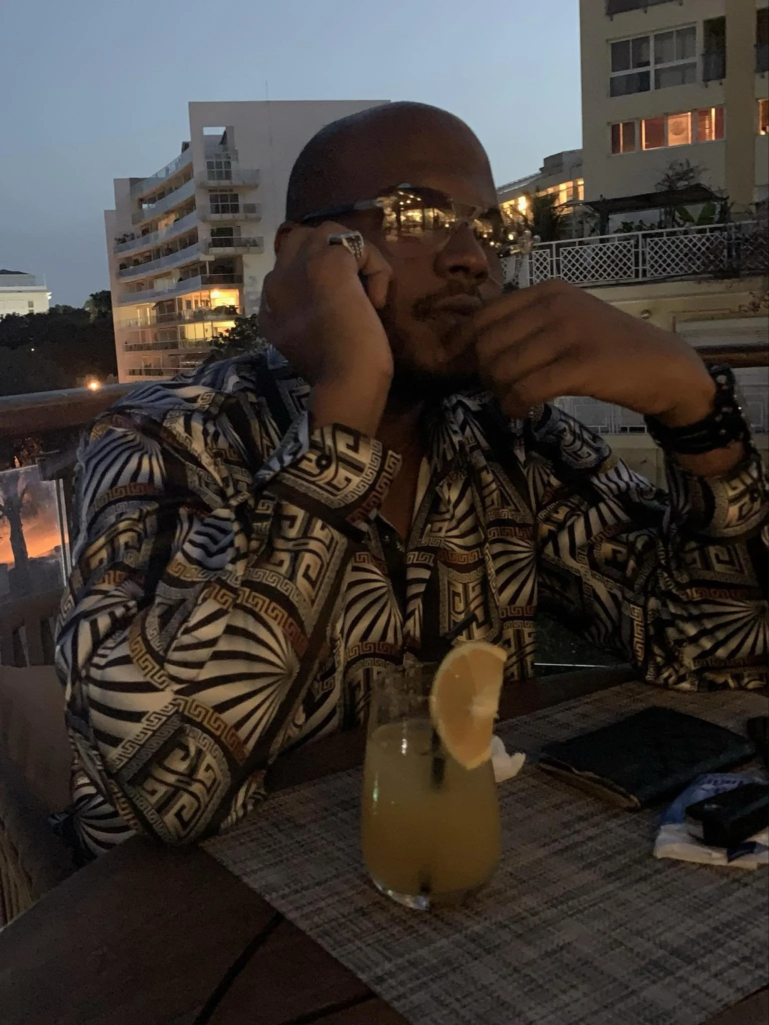 A man with a patterned shirt, wearing sunglasses, holding a phone to his ear, sitting at an outdoor restaurant table during dusk with a tropical cocktail decorated with a lemon wedge in the foreground.