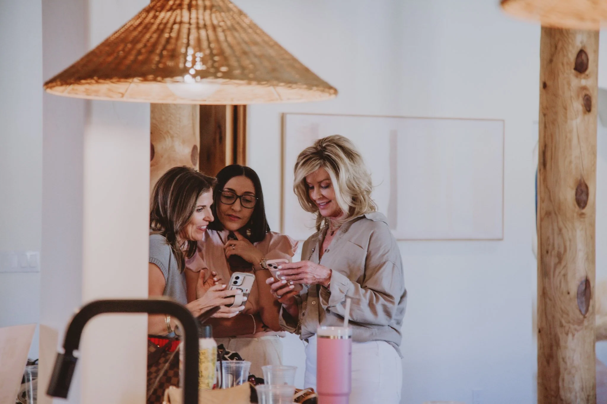 Three women gathered around sharing and looking at their phones in a cozy indoor setting, with a large hanging light and natural wooden elements.