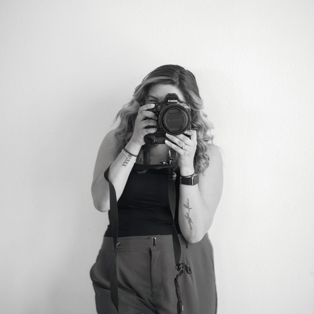 A woman with long, wavy hair taking a photo with a Canon camera in front of a plain wall.
