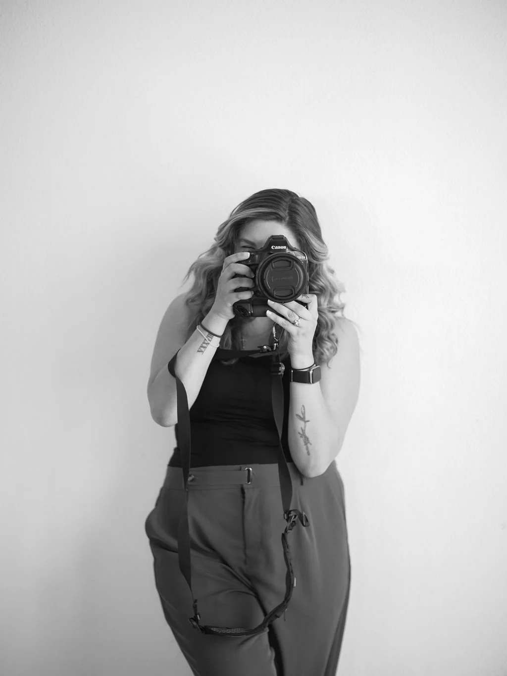 Black-and-white photo of a woman taking a selfie with a camera, standing against a plain wall. She has wavy hair, tattoos on her arms, and is wearing a sleeveless top and high-waisted pants.