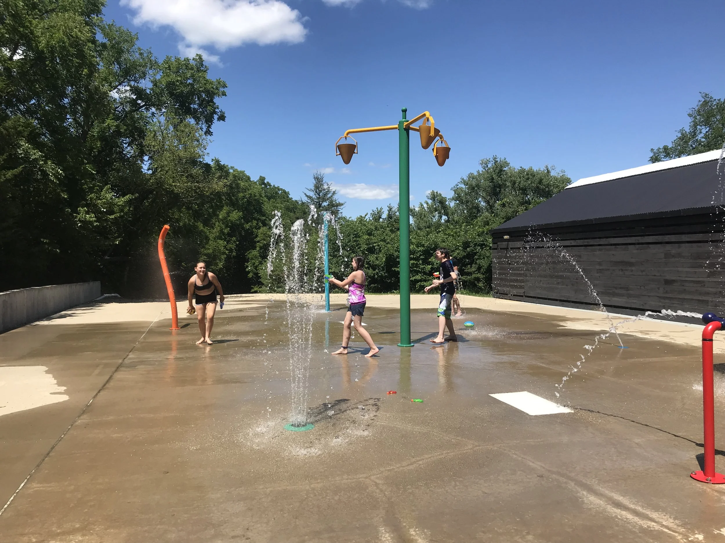 Children playing at a splash pad with water fountains and sprayers under a clear blue sky.