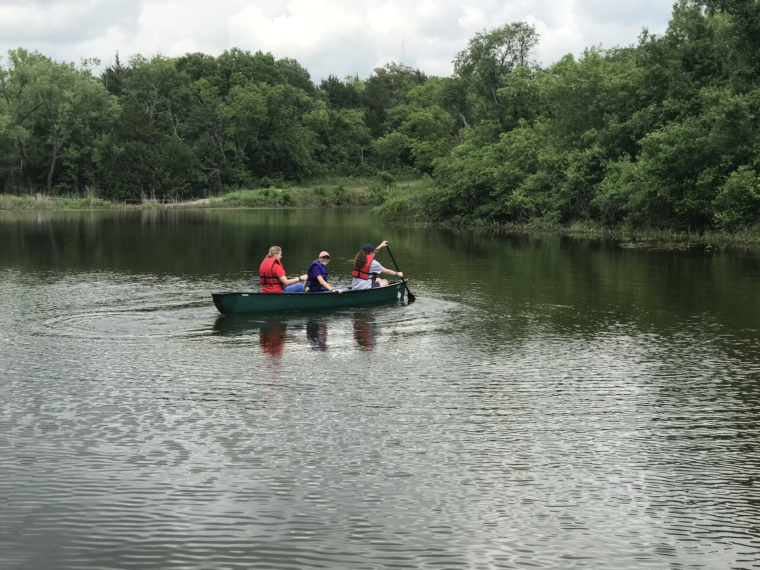Three people in a green canoe paddling on a calm lake surrounded by trees.