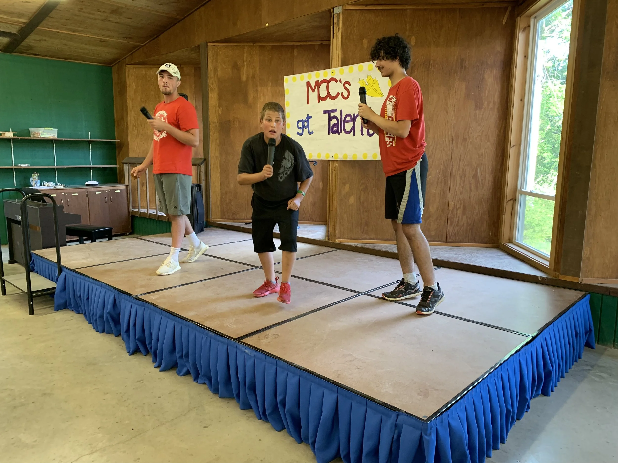 Three people holding microphones on a stage with a banner reading "MCC's got Talent."