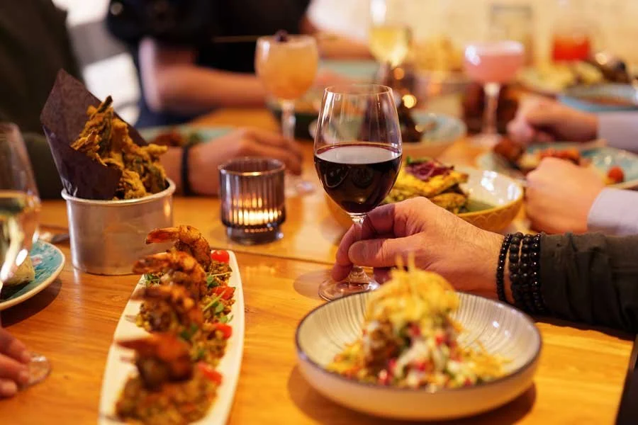 Focus on a glass of red wine surrounded by Pukka chaat, prawns and okra fries on a wooden table