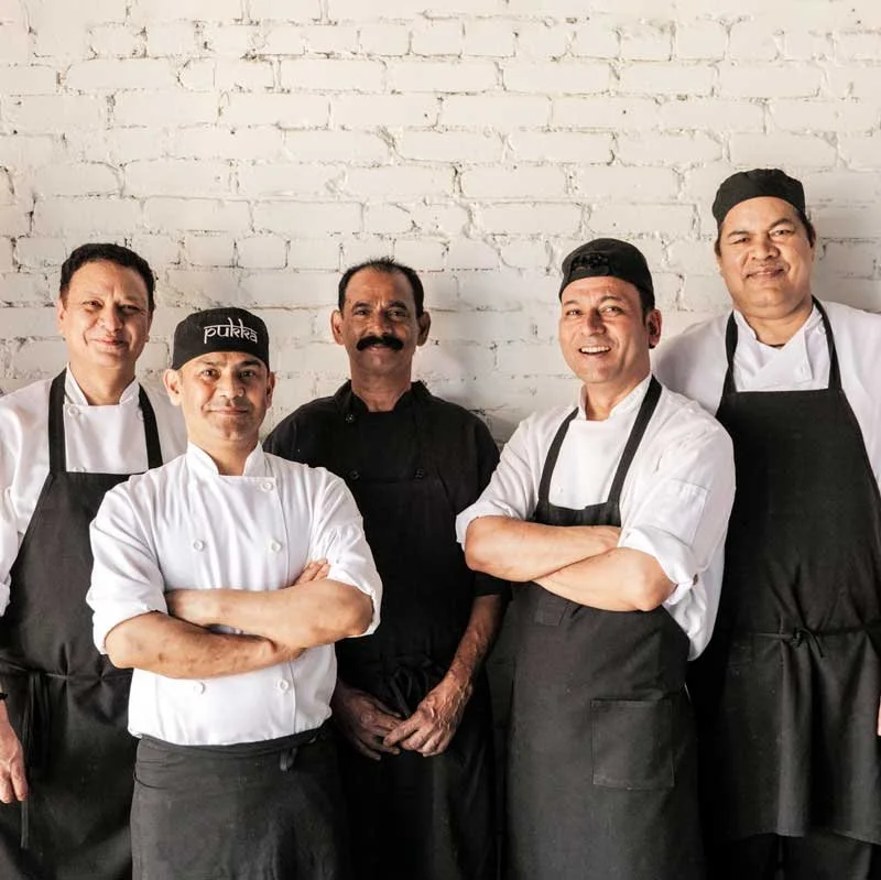 Group of five chefs standing against a white brick wall, wearing chef uniforms and aprons, smiling.