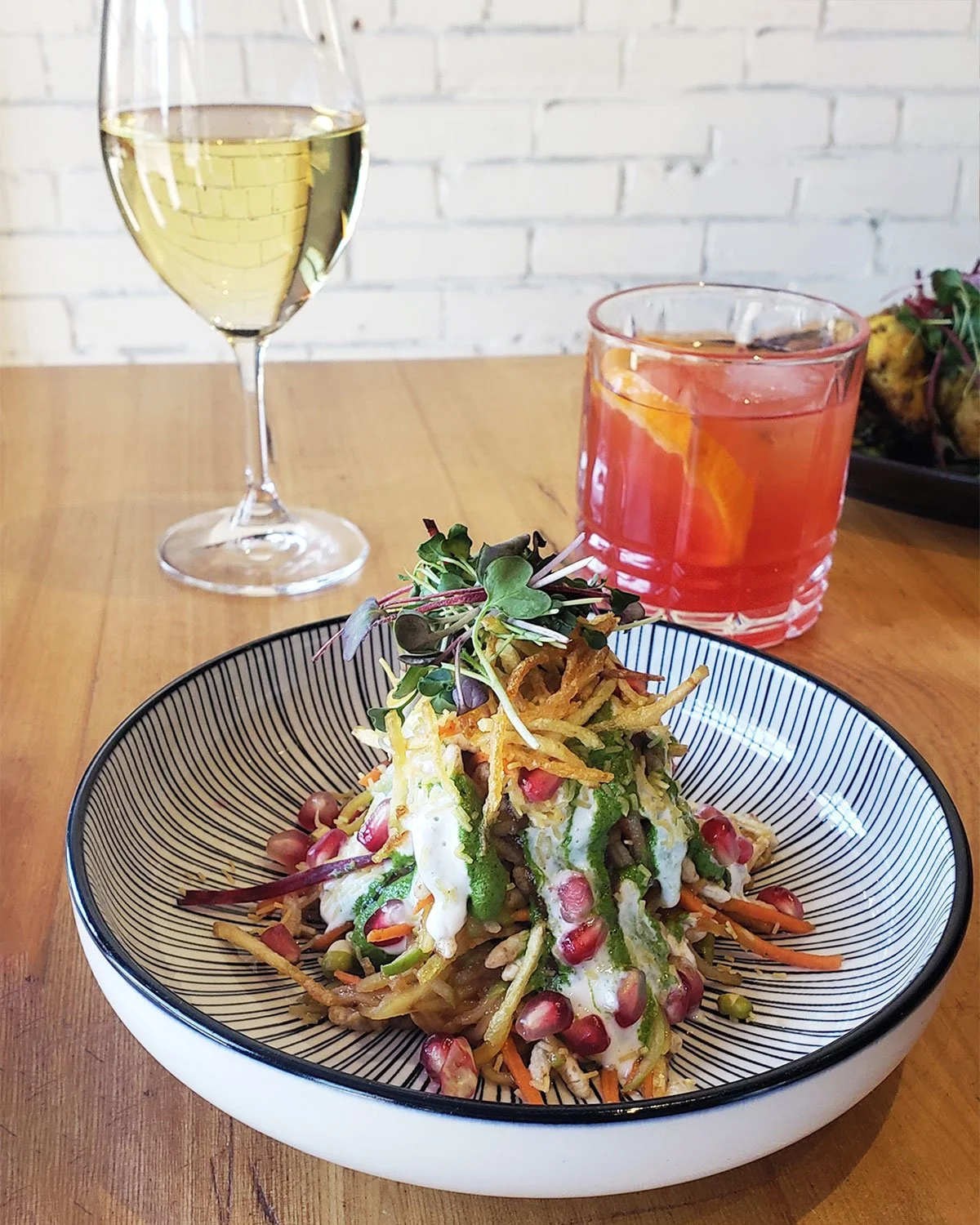 A plate of colorful chaatr with microgreens on top, served with a glass of white wine and a red cocktail on a wooden table, with a white brick wall in the background.