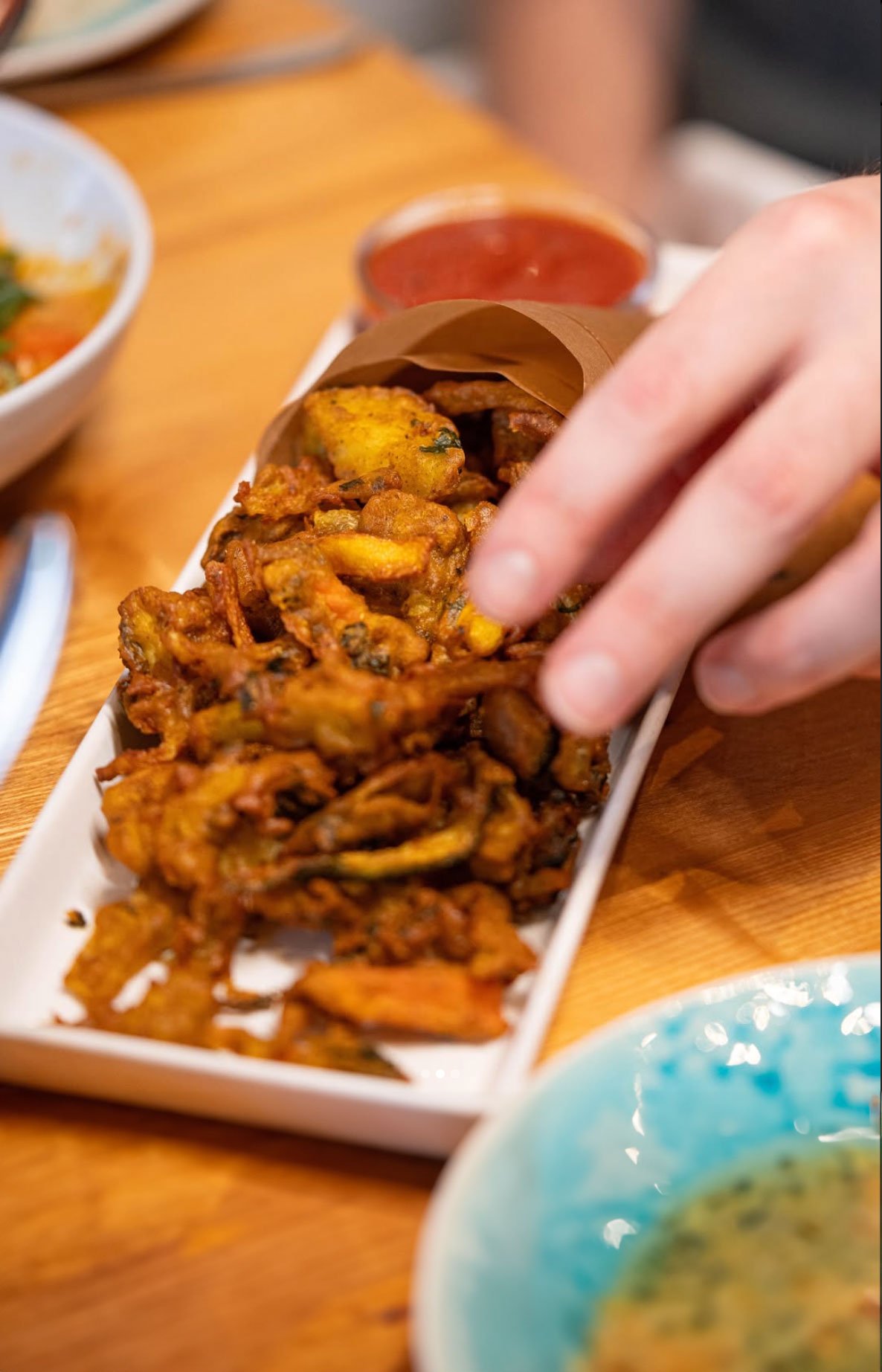 A hand reaching for Indian pakoras on a white serving dish, with a bowl of red dipping sauce behind them, on a wooden table.