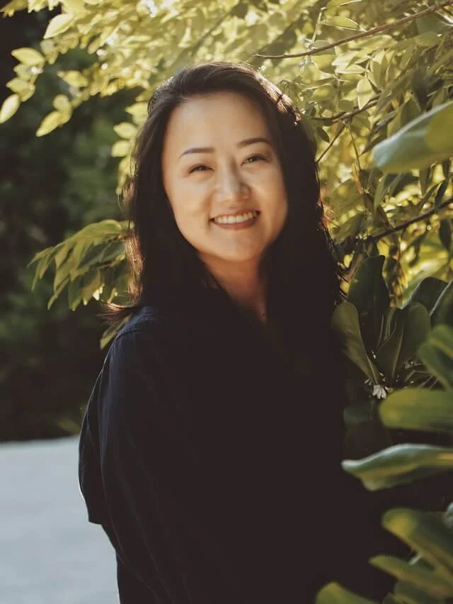 A smiling woman with dark hair standing outside with foilage in the background