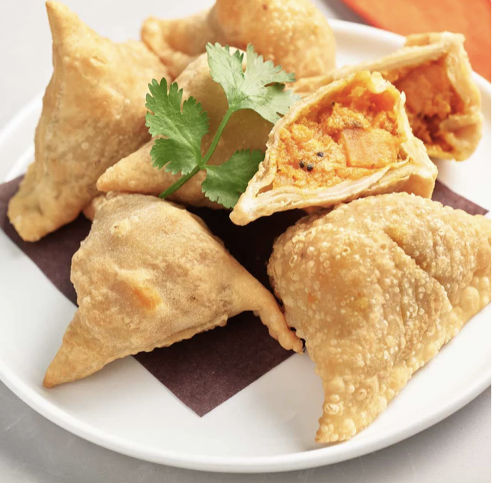 Assorted fried samosas with one cut in half revealing an orange vegetable filling, garnished with cilantro on a white plate.