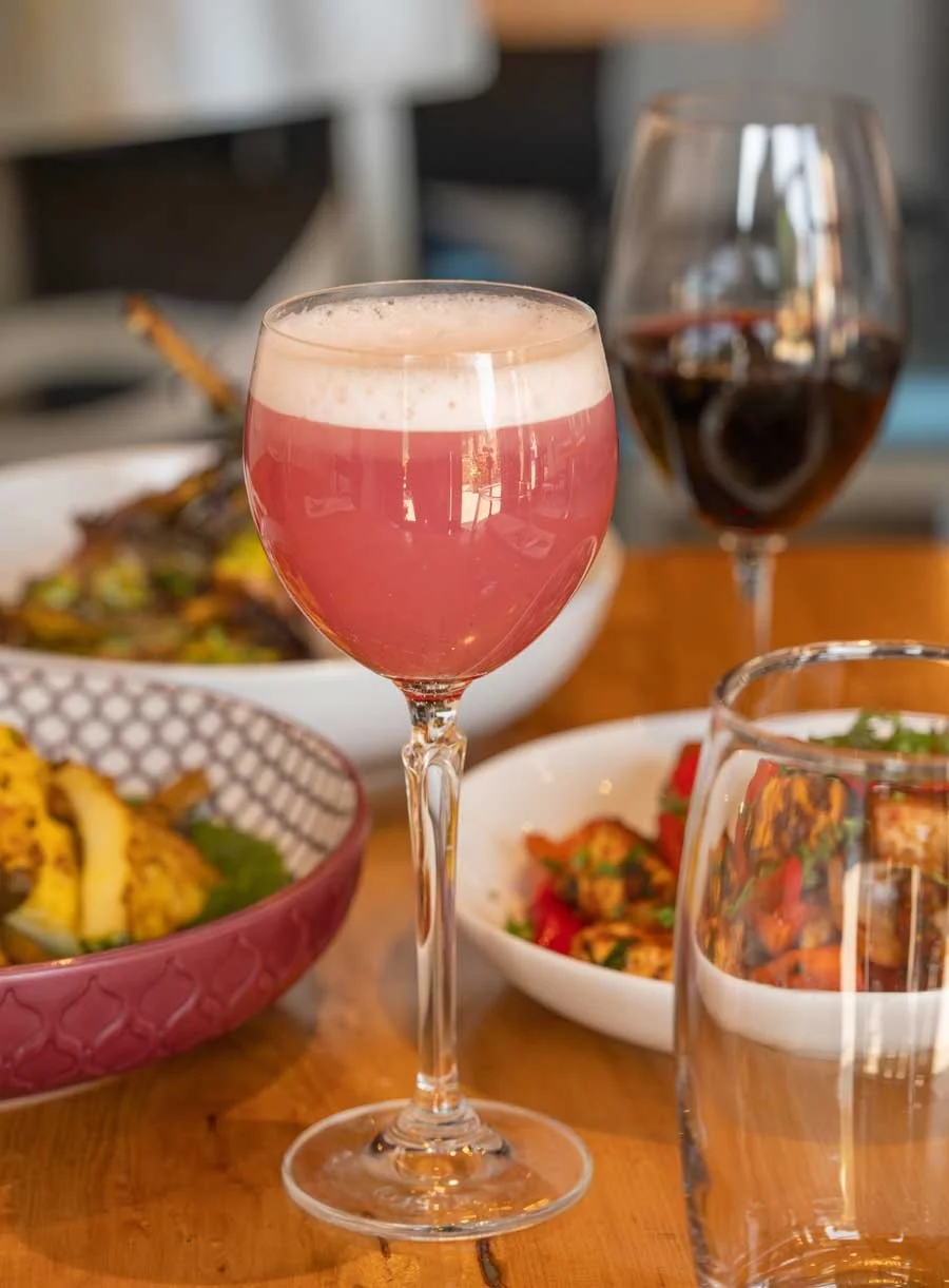 A pink cocktail in a tall stemmed glass surrounded by food