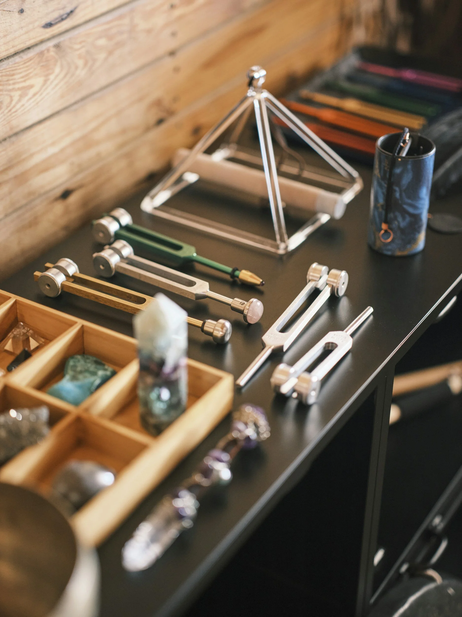 Various tools and craft supplies on a black table, including geometric metal and wooden frames, a container holding pens and pencils, a small wooden box with compartments, and colored pencils in a pouch, with a wooden wall in the background.