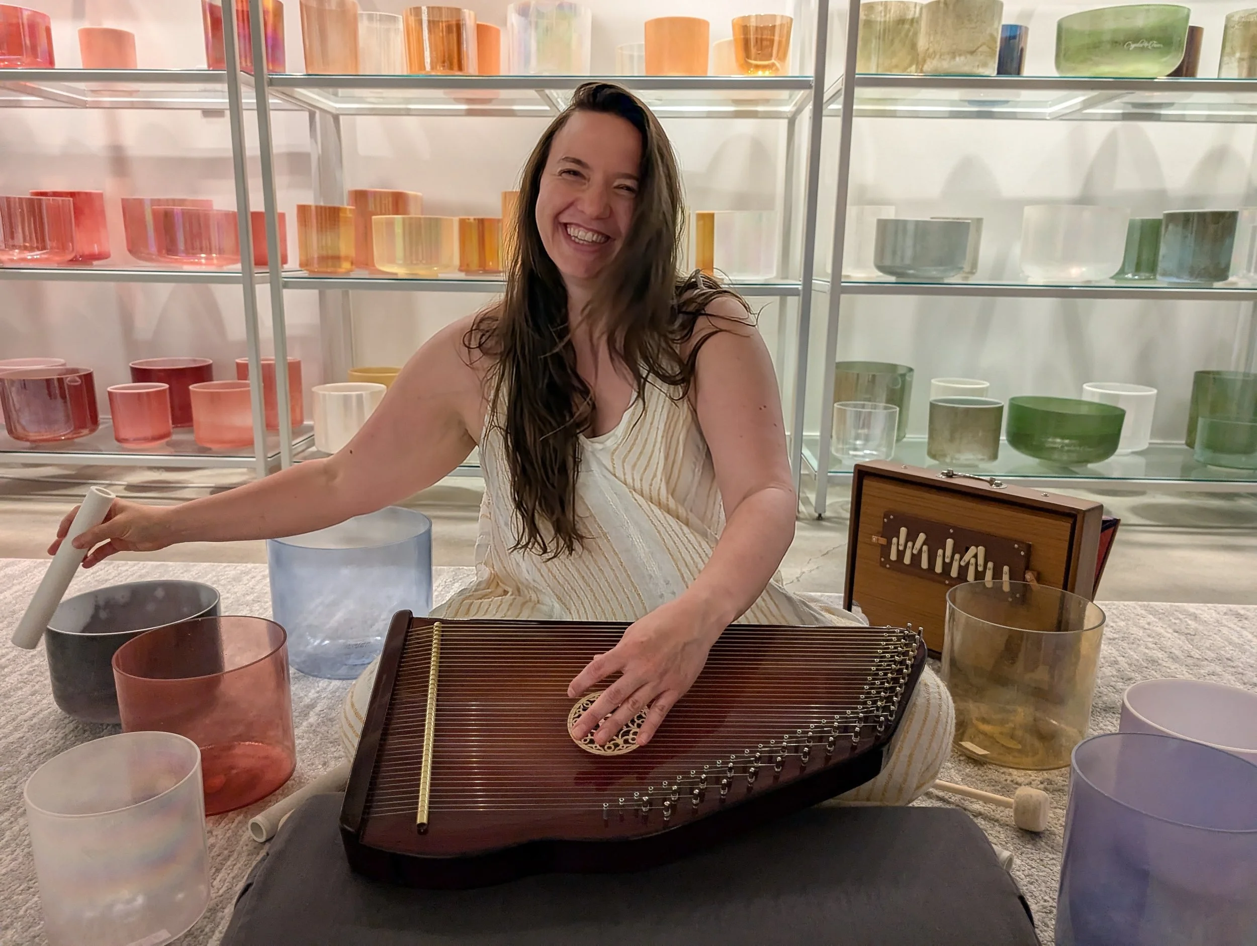 A woman with long dark hair smiling and playing crystal singing bowls and therapy harp, surrounded by colorful glass bowls on shelves. She is sitting on a carpeted floor with other musical instruments and glassware nearby.