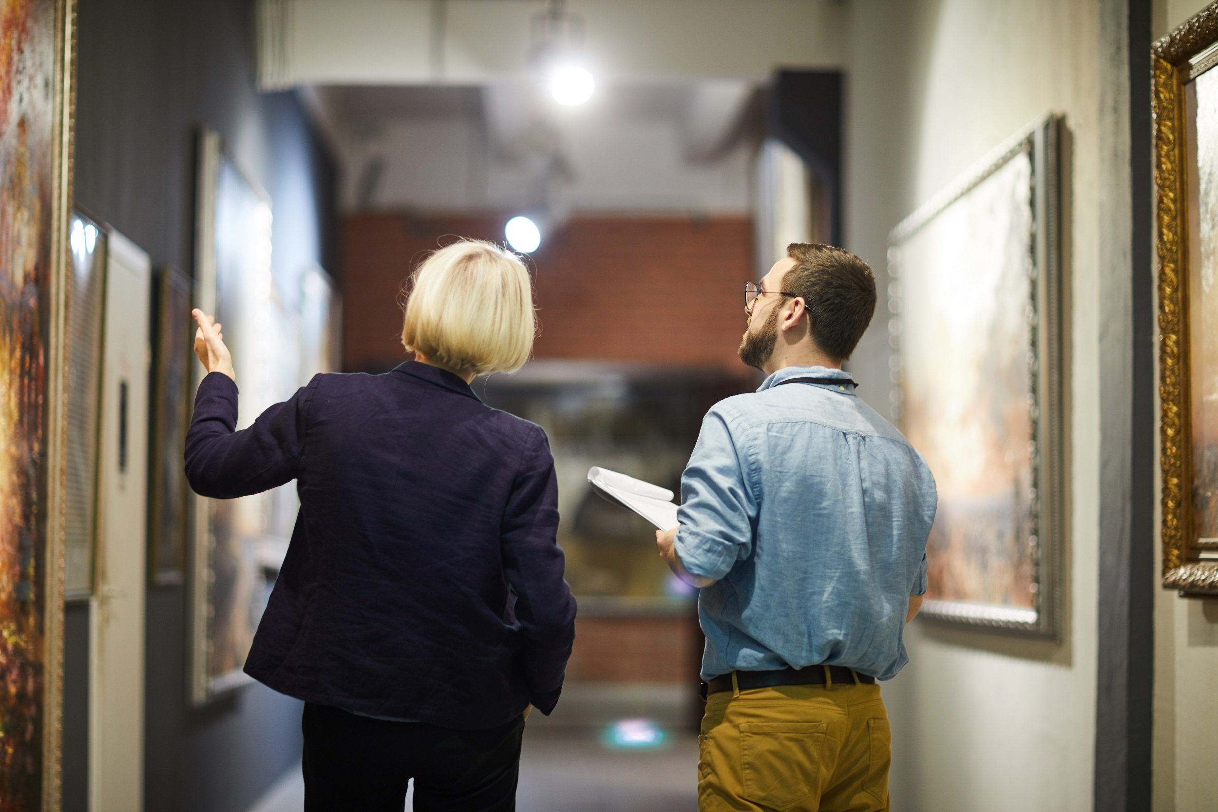 Back view portrait of two museum workers discussing paintings walking in art gallery