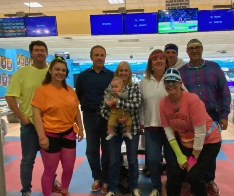 RKM brokers dressed in 80s gear at a bowling alley. Bowling screens and lanes are visible in the background. Logos for Big Brothers Big Sisters of Canada and RKM are present in the image.