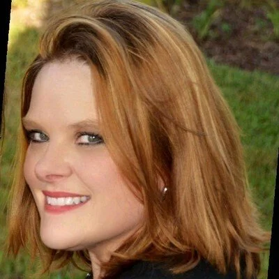 A young woman with shoulder-length red hair, smiling outdoors in a grassy area.