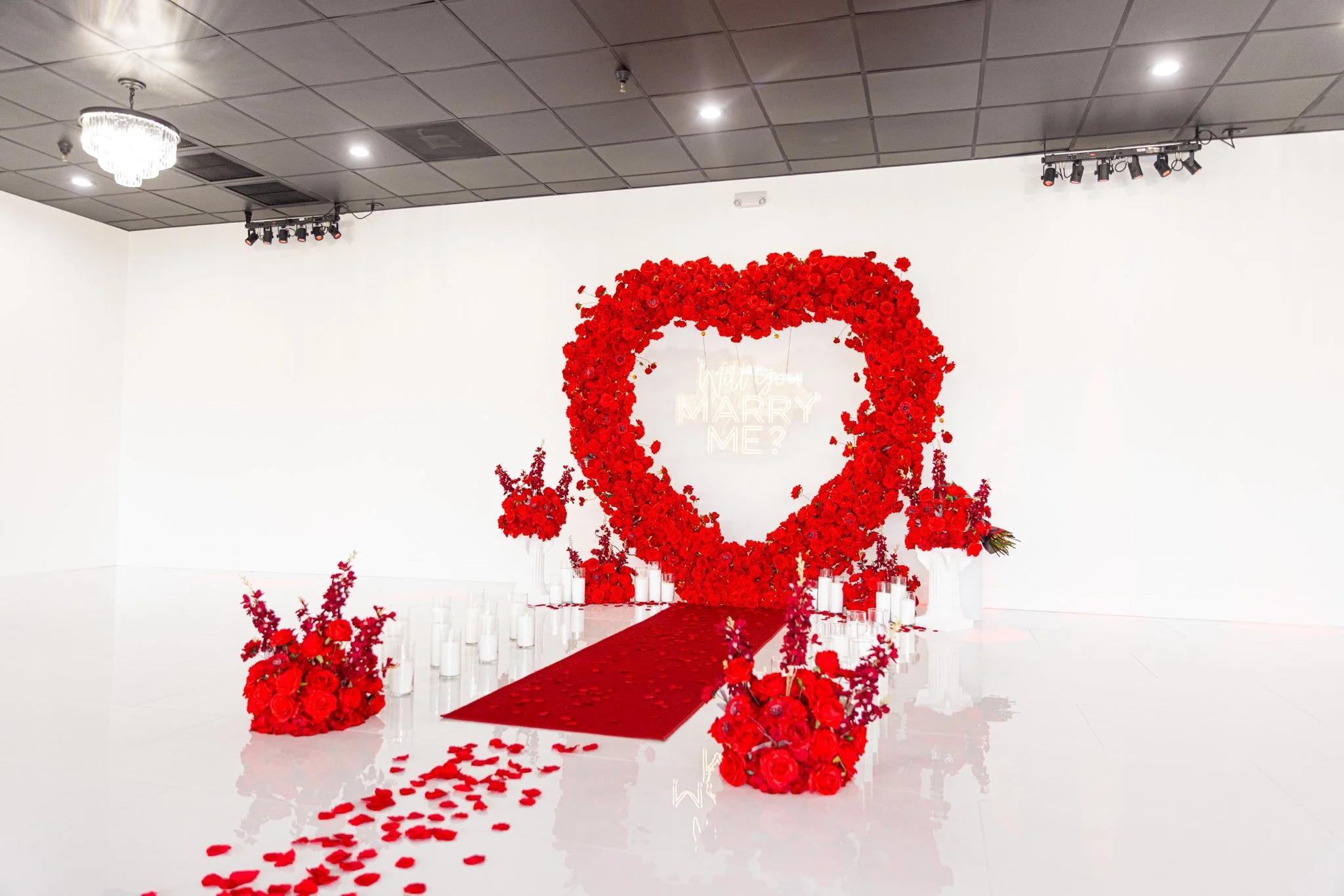  Beautiful heart-shaped proposal arrangement of red roses, rose pedals, white candles, and red carpet in our stunning event venue. Decor Designed and Executed by  Orlando Elite Decor  