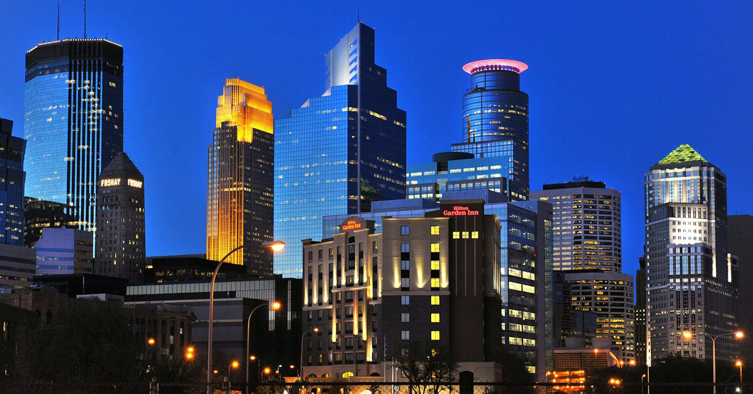 Nighttime skyline of high-rise buildings in downtown, illuminated with colorful lights against a dark blue sky.
