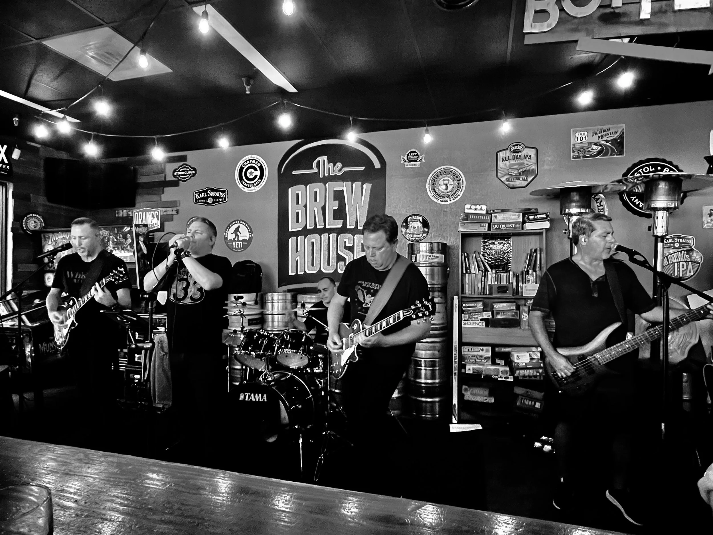 A band performing live music in a bar with a sign on the wall that says 'The Brew House', overhead string lights, and shelves with books and signs.
