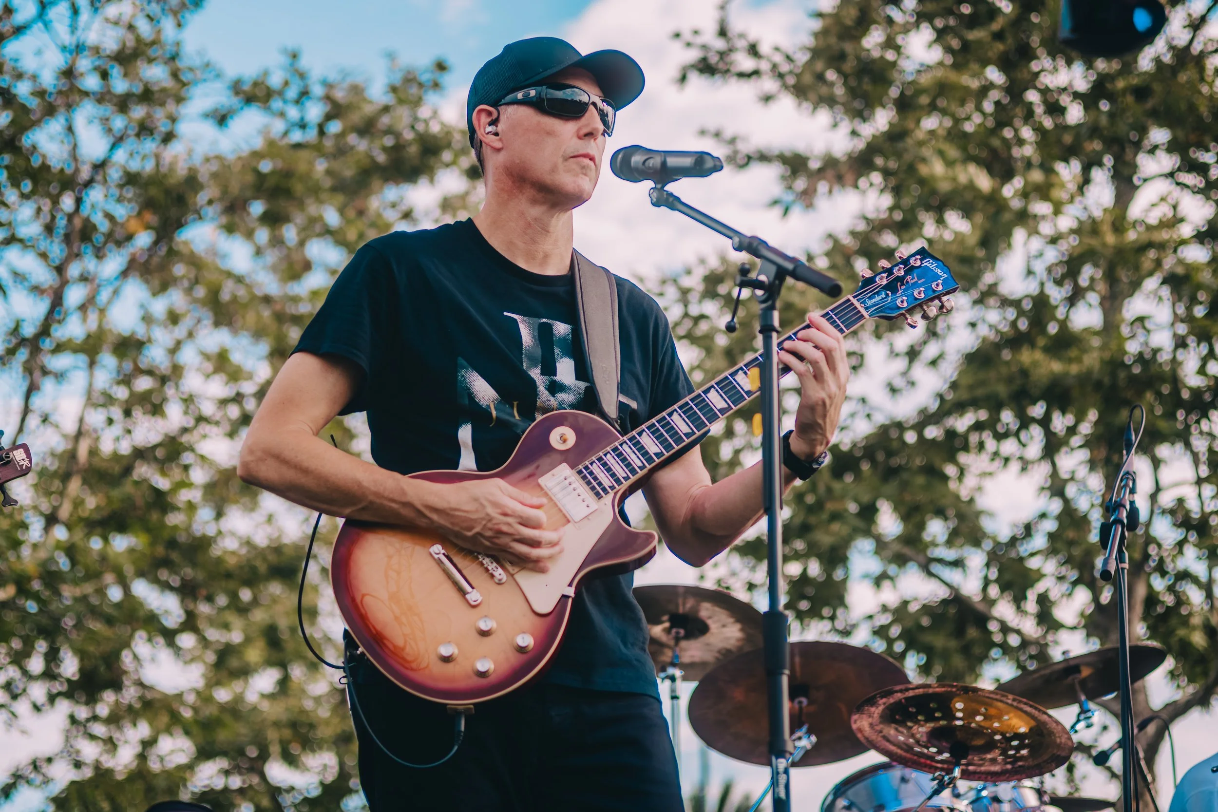 A man wearing sunglasses, a cap, and a black T-shirt playing an electric guitar outdoors during the daytime, with trees and a drum set visible in the background.