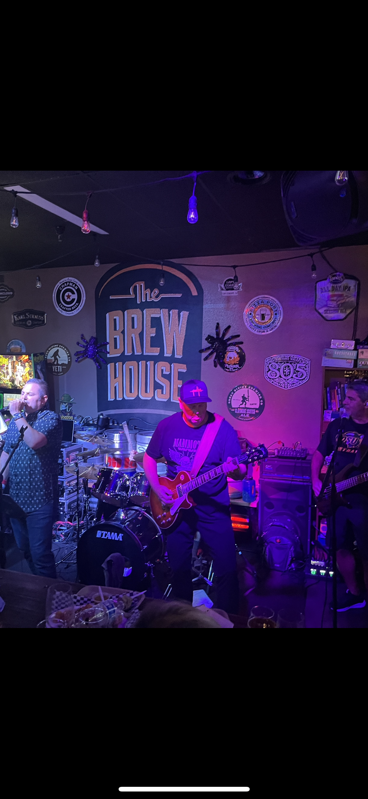 Live band performing at The Brew House, with musicians playing guitar and drums, decorated with beer and brewery-themed signs and lights on the wall.