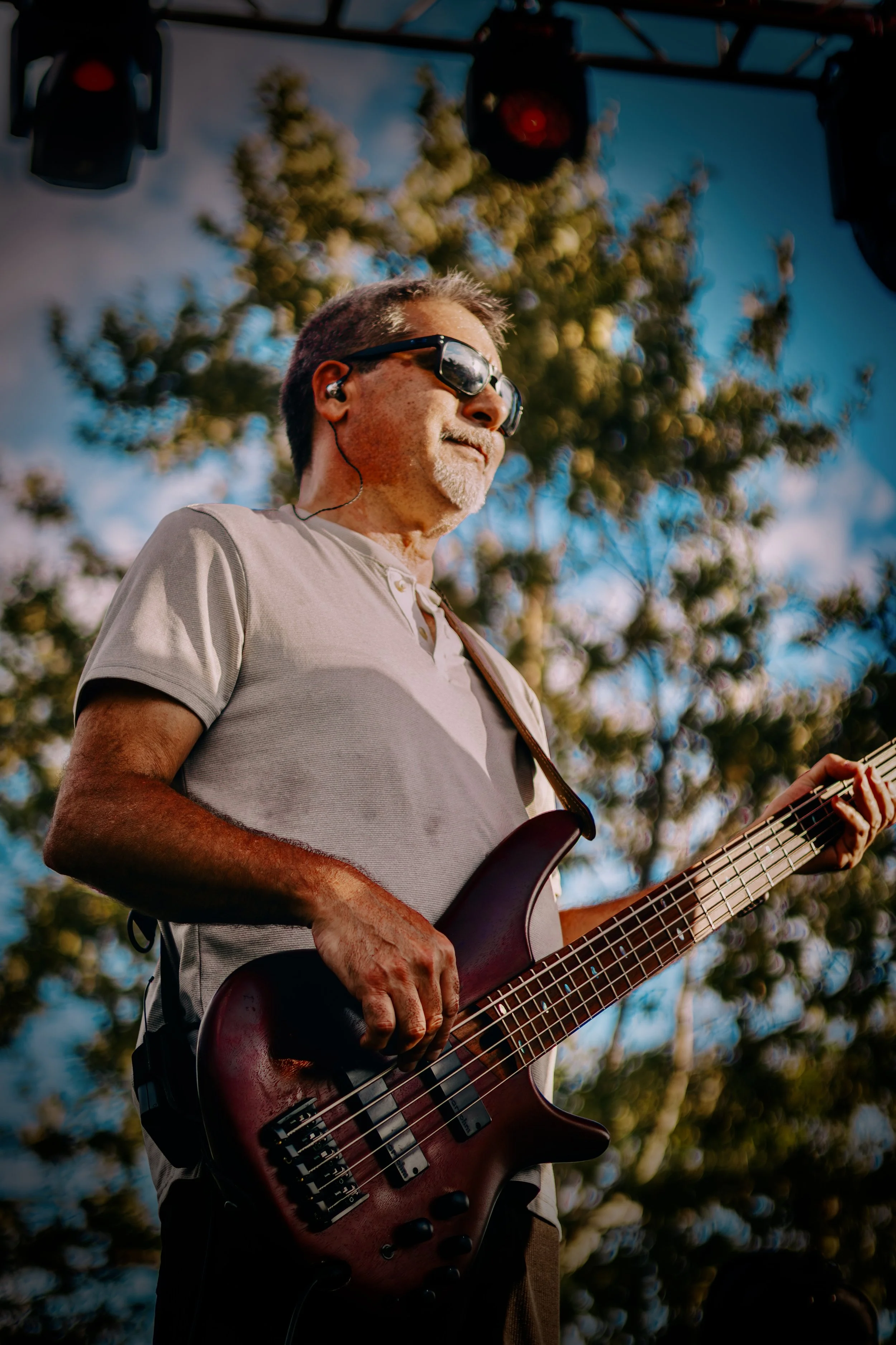 A man playing an electric bass guitar outdoors under a tree, wearing sunglasses and earphones, during a sunny day.