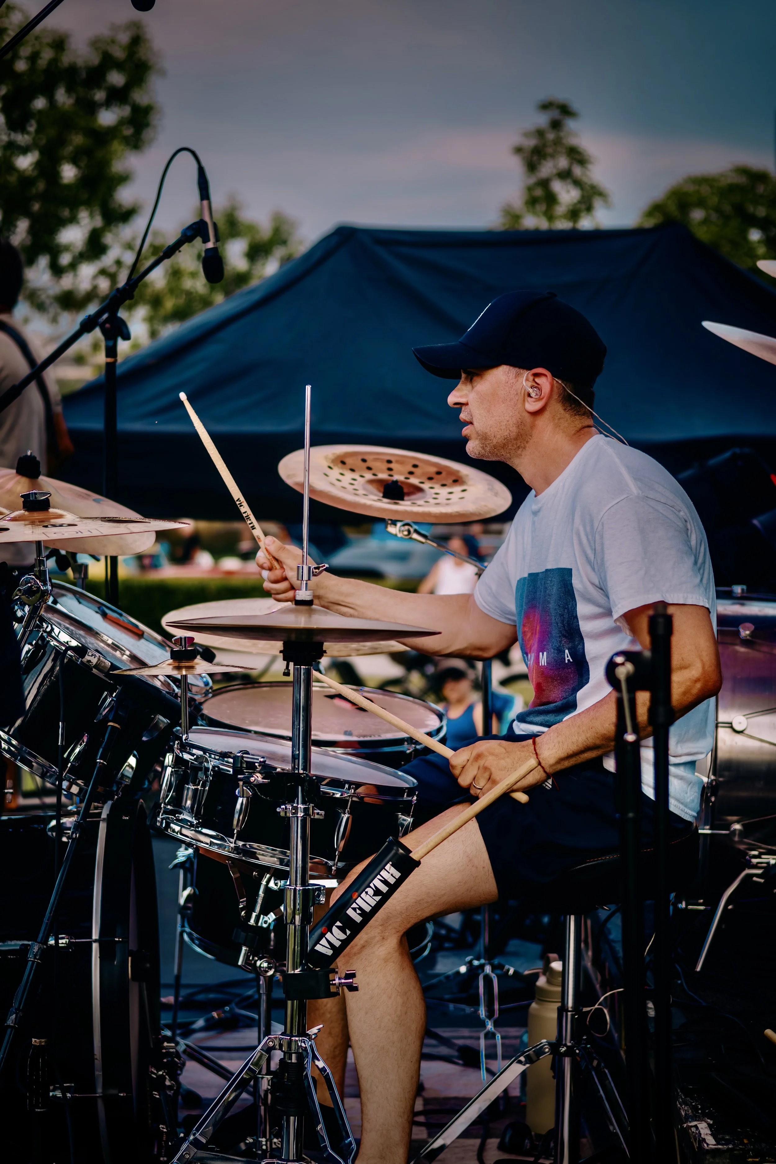 A man wearing a black cap, white T-shirt, and black shorts is playing the drums outdoors. He is focused and holding drumsticks while sitting behind a drum set, with a large blue tent and trees in the background.