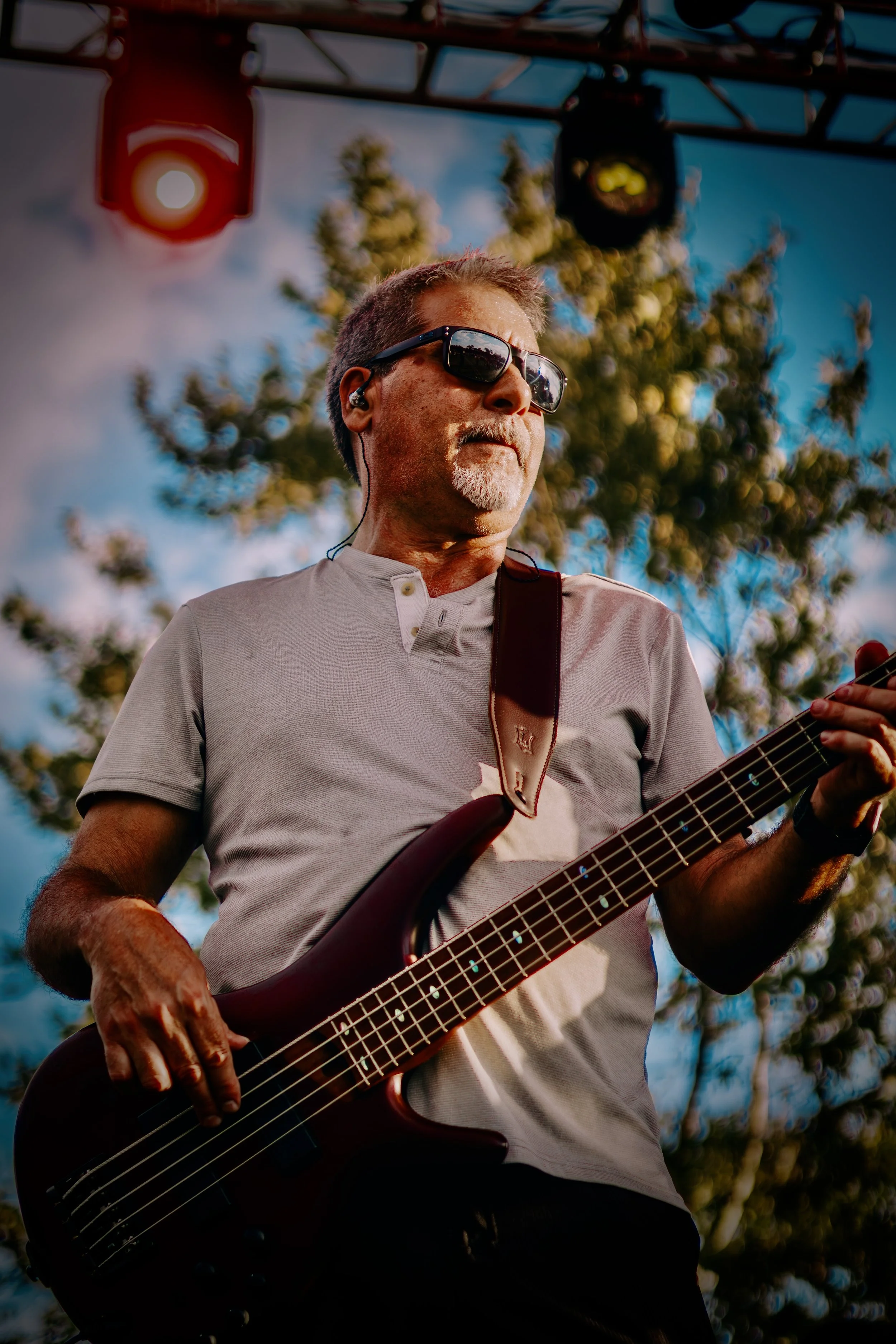 A man playing a bass guitar outdoors during sunset, wearing sunglasses, a gray shirt, and headphones, with trees and a blue sky in the background.